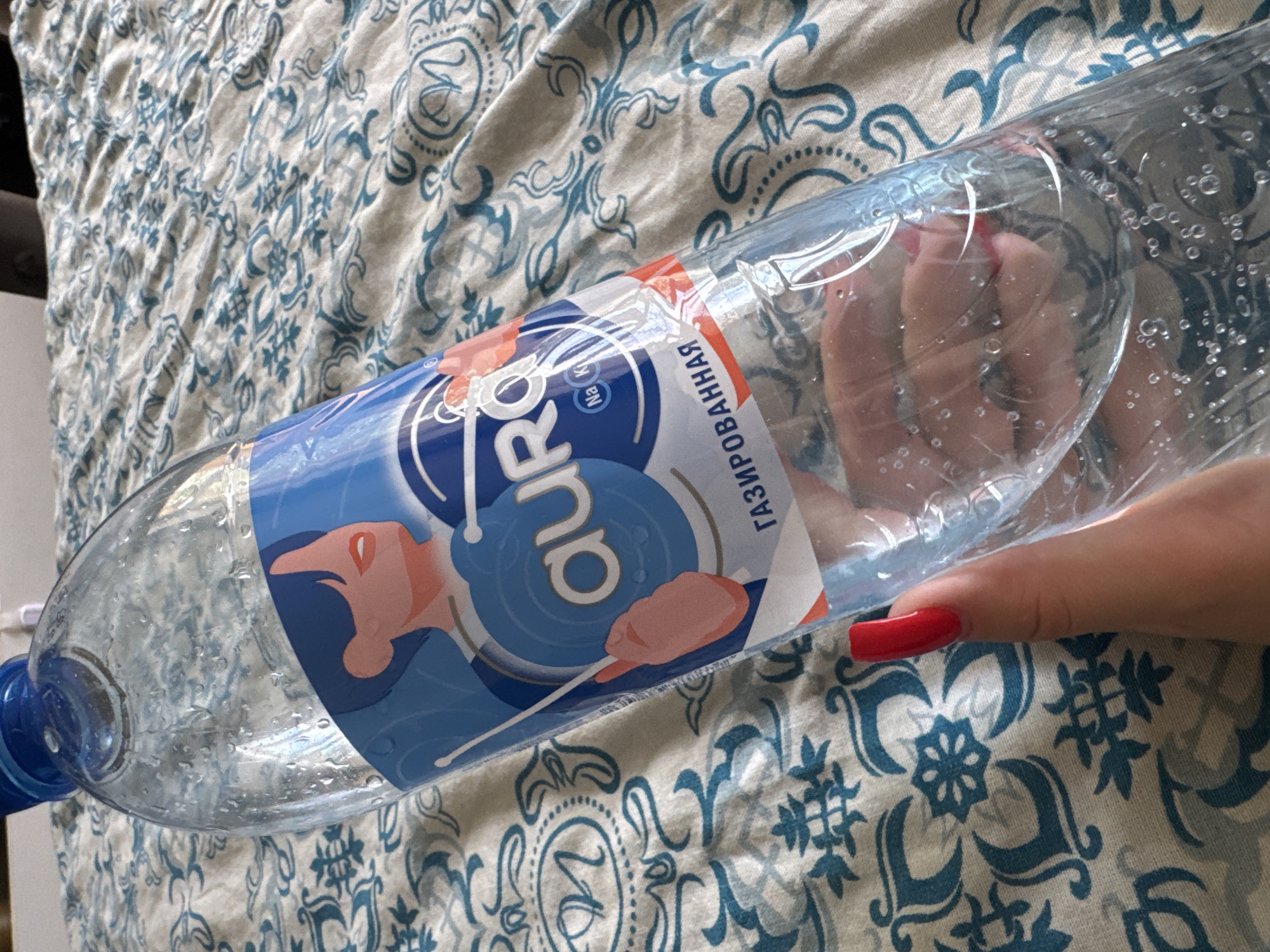 A clear plastic bottle of carbonated water, labeled "Aura", is held by a person with bright red fingernails against a background of a blue and white patterned fabric. The bottle is partially full and condensation is visible on its surface. The label features a stylized illustration of a person's face and the text "газированная" (carbonated in Russian). The scene appears to be indoors, likely on a bed or couch. The location is described as Яромінскі сельскі Савет, Belarus, though this cannot be verified from the image itself.