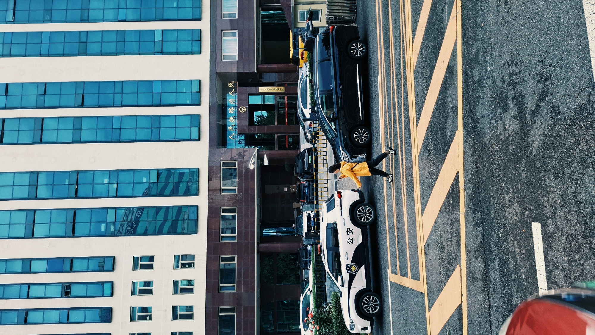 The image shows a street scene in the Dongcheng District, China. A modern building with horizontal rows of blue-tinted windows stands next to a building with dark brown facade and signage with Chinese characters. A person wearing a yellow jacket is walking across a street marked with yellow diagonal stripes, between parked cars. The vehicles include a white police car marked with Chinese characters and black vehicles.