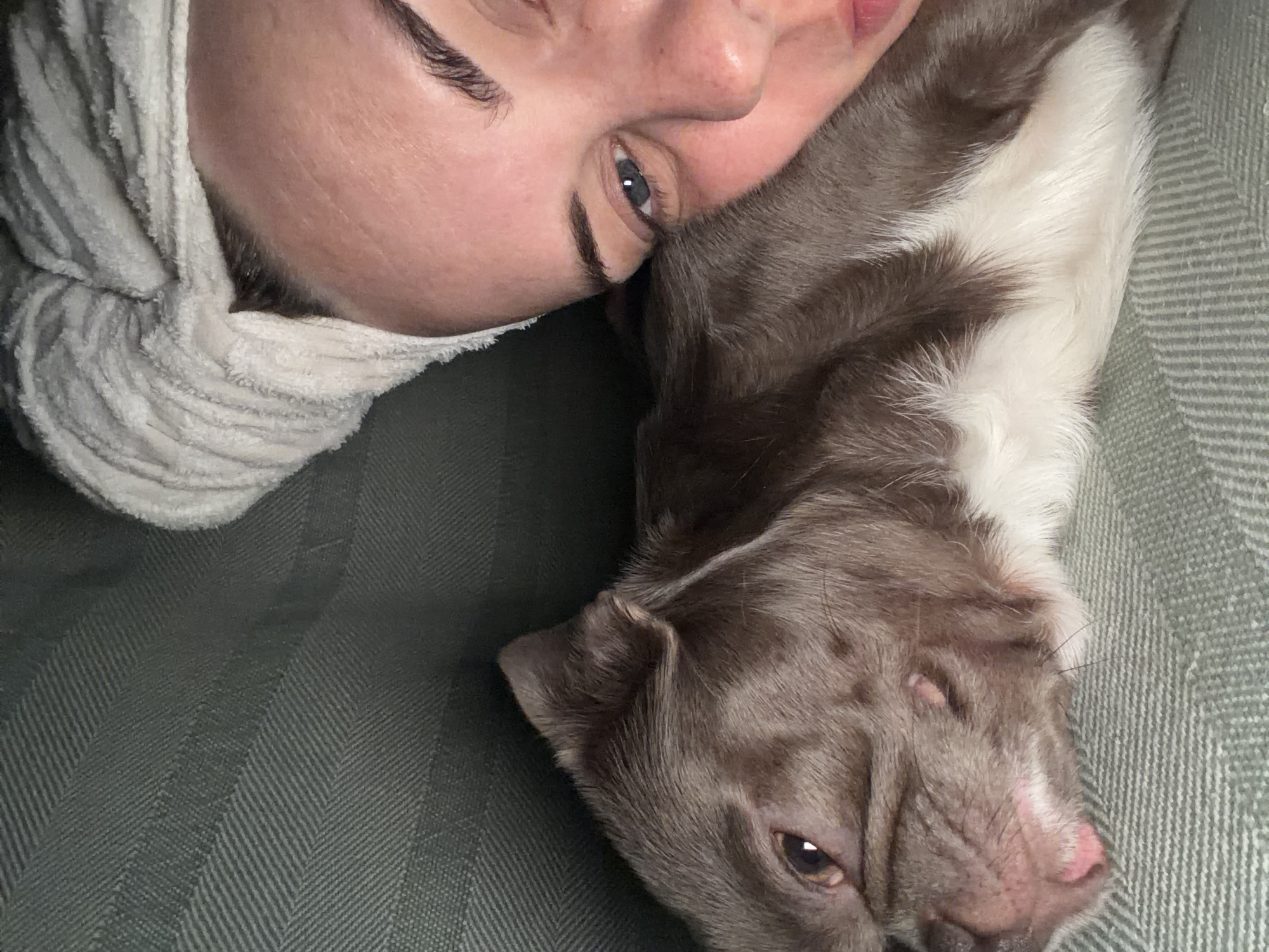 A person is lying down next to a dog. The person has light skin, dark eyebrows, and blue eyes. They are wearing a white towel wrapped around their head. The dog is primarily brown with some white fur. Both are lying on a green herringbone-patterned surface. There is no indication of the location being Votkinsk, Russia.