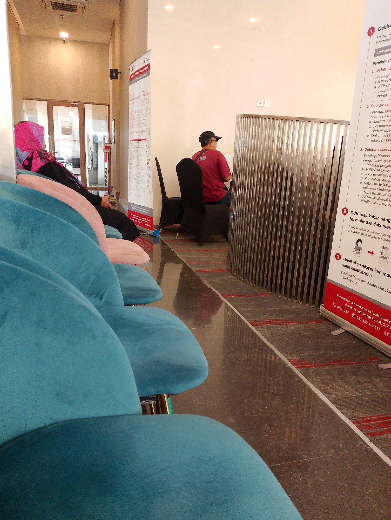 An indoor waiting area in what appears to be a government office or financial institution. The floor is polished concrete with red striped carpet markers. A row of plush teal and pink chairs is arranged in the foreground. In the mid-ground, a person wearing a red shirt and a black cap sits on a black chair. To the right of this person, a metal partition with vertical slats separates the waiting area from another space. A large white banner with red lettering is displayed on a stand to the far right, featuring information about debitur requirements and OJK (Financial Services Authority) procedures. A woman wearing a pink hijab is seated in the background to the left, partially obscured by the chairs. The lighting is bright, suggesting overhead fluorescent lights. The environment is clean and orderly.