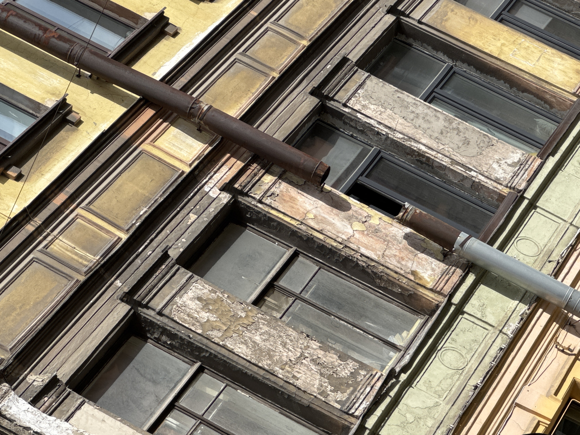 The image presents an upward, angled view of a building facade in Saint Petersburg, Russia. The facade exhibits architectural details of multiple windows with frames in varying states of disrepair. The paint on the window frames is peeling and worn, suggesting exposure to the elements. A rusty pipe runs diagonally across the facade, obscuring portions of the windows and the building's surface. The wall colors range from yellow to beige to light green. The building's design features decorative elements. The windows appear to be closed. The sky is not visible.