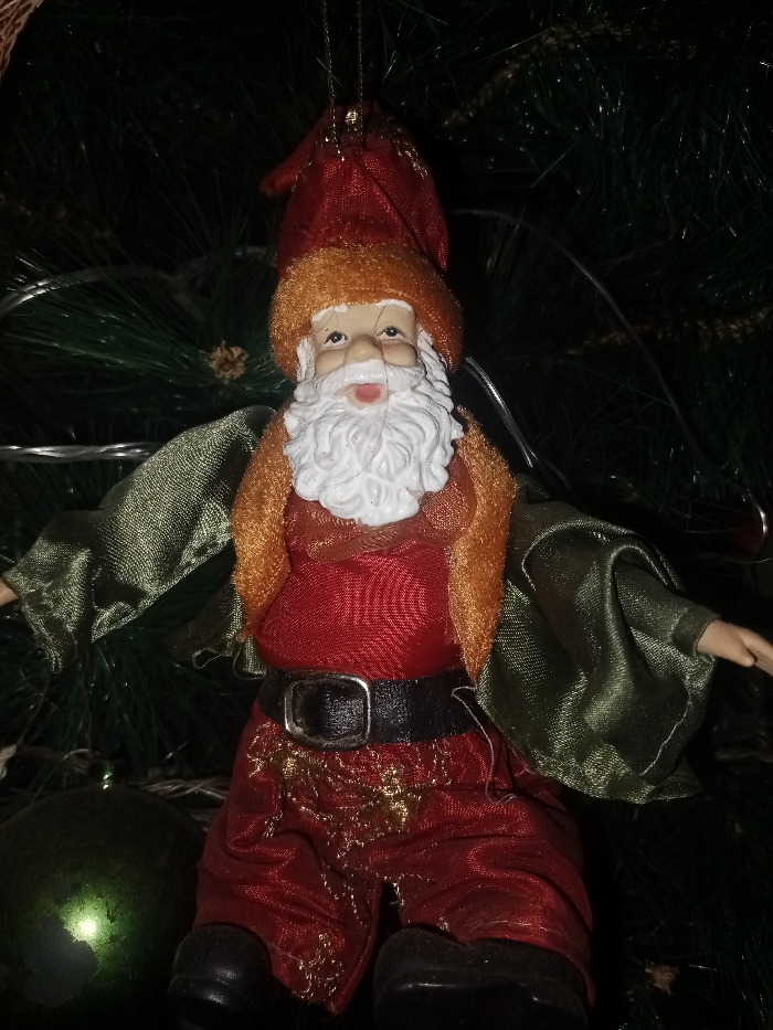 A Christmas tree ornament depicting a Santa Claus-like figure. The doll wears a red cap, red shirt, and red pants with gold embellishments, secured by a black belt with a silver buckle. It also has a golden collar and green sleeves that stretch out to the sides. It features a white beard and pale skin tone. A dark green Christmas bulb is visible in the lower left of the frame. The entire scene is set against a background of green Christmas tree branches and white fairy lights.