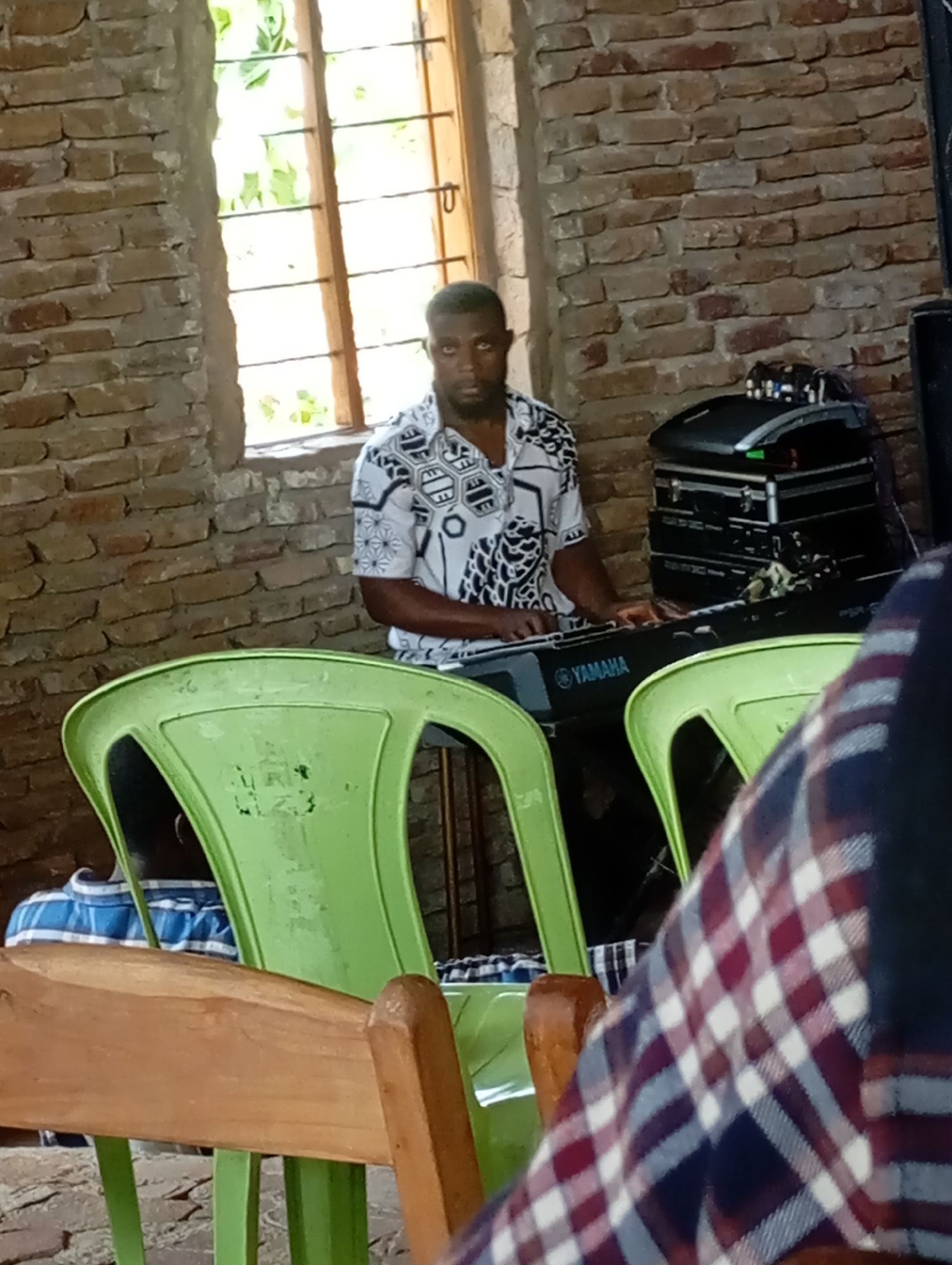 A man is sitting at a Yamaha keyboard inside what appears to be a brick-walled building in Uke, Nigeria. Behind him, a window with a view of greenery lets in natural light. To his right is a black audio equipment rack. In the foreground, a light green plastic chair is visible along with parts of other chairs occupied by people. The man playing the keyboard is wearing a short-sleeved patterned shirt.