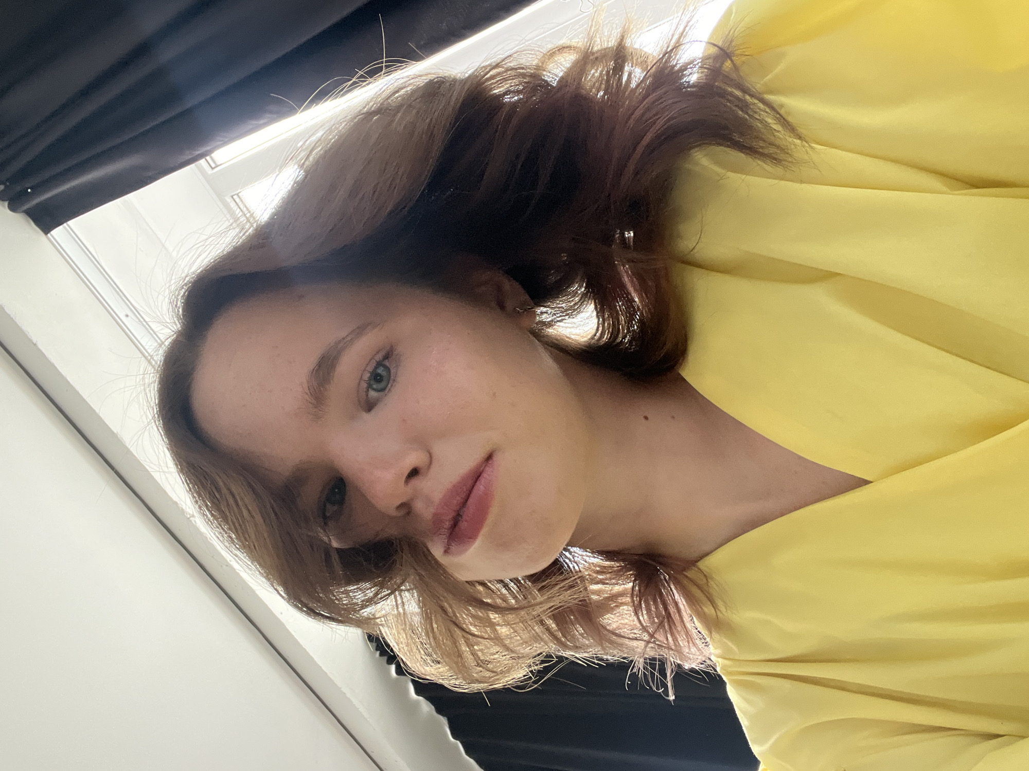 A woman is angled downward towards the camera, wearing a yellow garment. Her hair is light brown with darker roots. She has fair skin and blue eyes. In the background, there are dark curtains and a white wall or window frame. The image does not provide any information to determine if it was taken in Chaikovsky, Russia.
