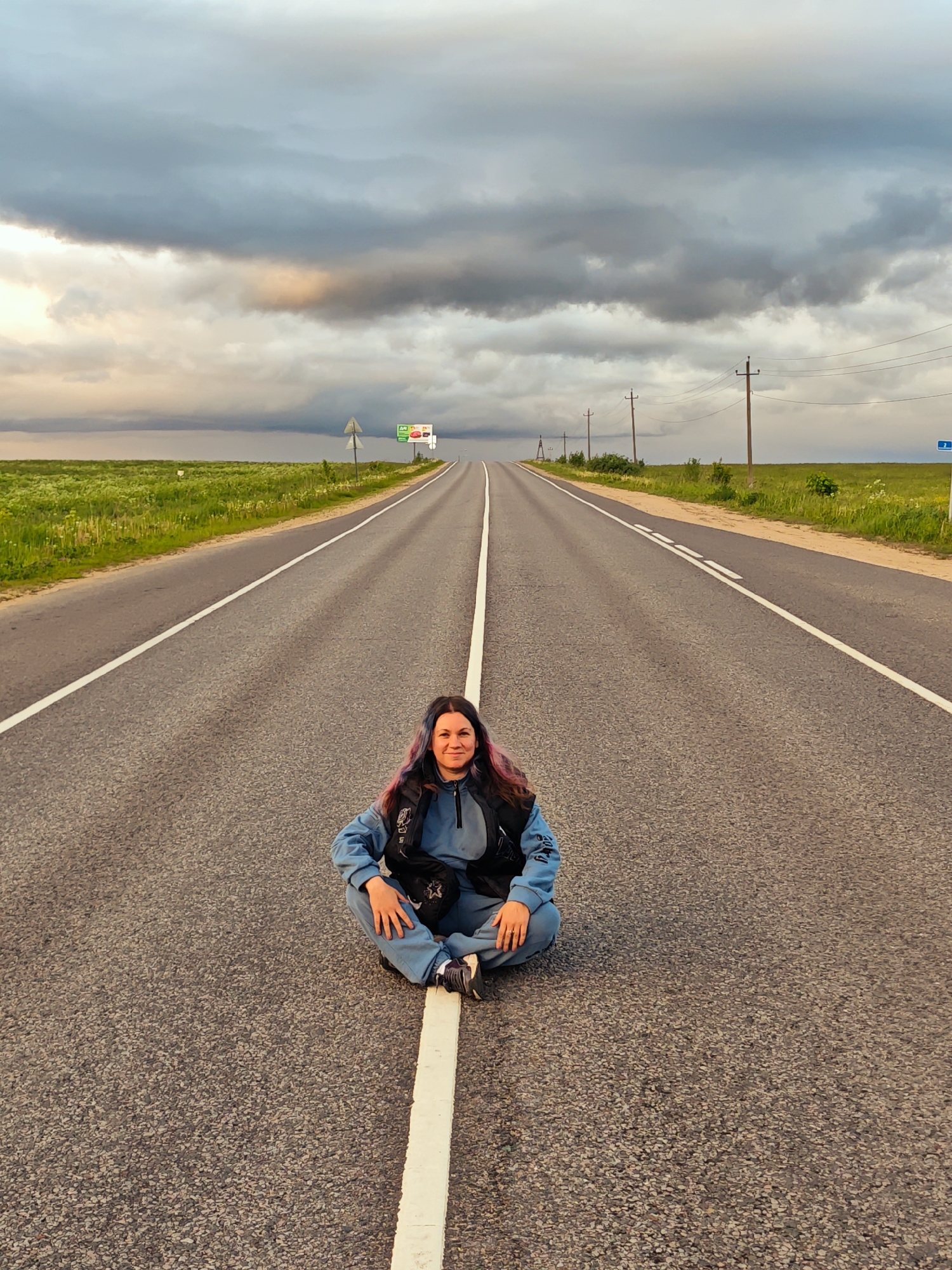 A medium shot depicts a woman sitting cross-legged on the white center line of a two-lane asphalt road. The location is near Курово, Russia. She is wearing a blue jumpsuit, a black vest, and dark sneakers. Her hair is long and dark with streaks of pink. The road stretches into the distance, under a cloudy sky with a patch of light near the horizon. Green fields border the road on both sides. Utility poles with wires run along the right side of the road. A billboard is visible in the distance on the left side of the road, along with some road signs.