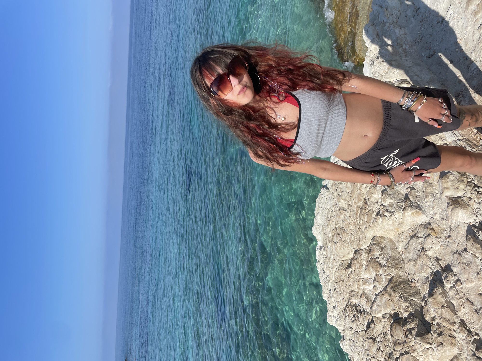 A young woman with long, wavy auburn and dark brown hair, wearing large brown sunglasses, a grey crop top, and black athletic shorts, stands on a rocky shoreline. The ocean, a vibrant turquoise, stretches out behind her under a clear blue sky. She is adorned with numerous bracelets and rings on her left wrist and fingers. The location is the coastline near Peyia, Cyprus, characterized by its rugged, light-colored rock formations and the crystal-clear waters of the Mediterranean Sea.