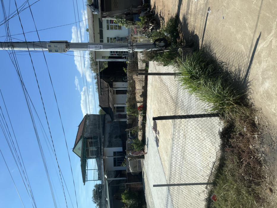 A residential scene showcases a power pole and multiple wires against a bright blue sky. Houses with varied architectural styles are scattered around the scene. A chain-link fence runs along the street, partially obscured by patches of dense grass and vegetation. A concrete sidewalk extends past the fence, with a dark shadow cast on it. The photo is taken outdoors on a sunny day.