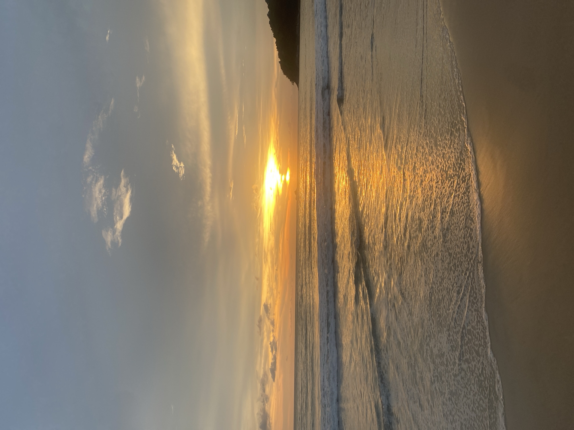 The image depicts a sunset at a beach in Choeng Thale, Thailand. The sun is low on the horizon, partially obscured by clouds, casting a golden glow across the sky and reflecting on the water. A dark headland is visible on the right side of the frame. Waves are gently breaking on the shore, creating a foamy edge between the water and the sand. The sand appears to be a light golden color. There are no people visible in the image.
