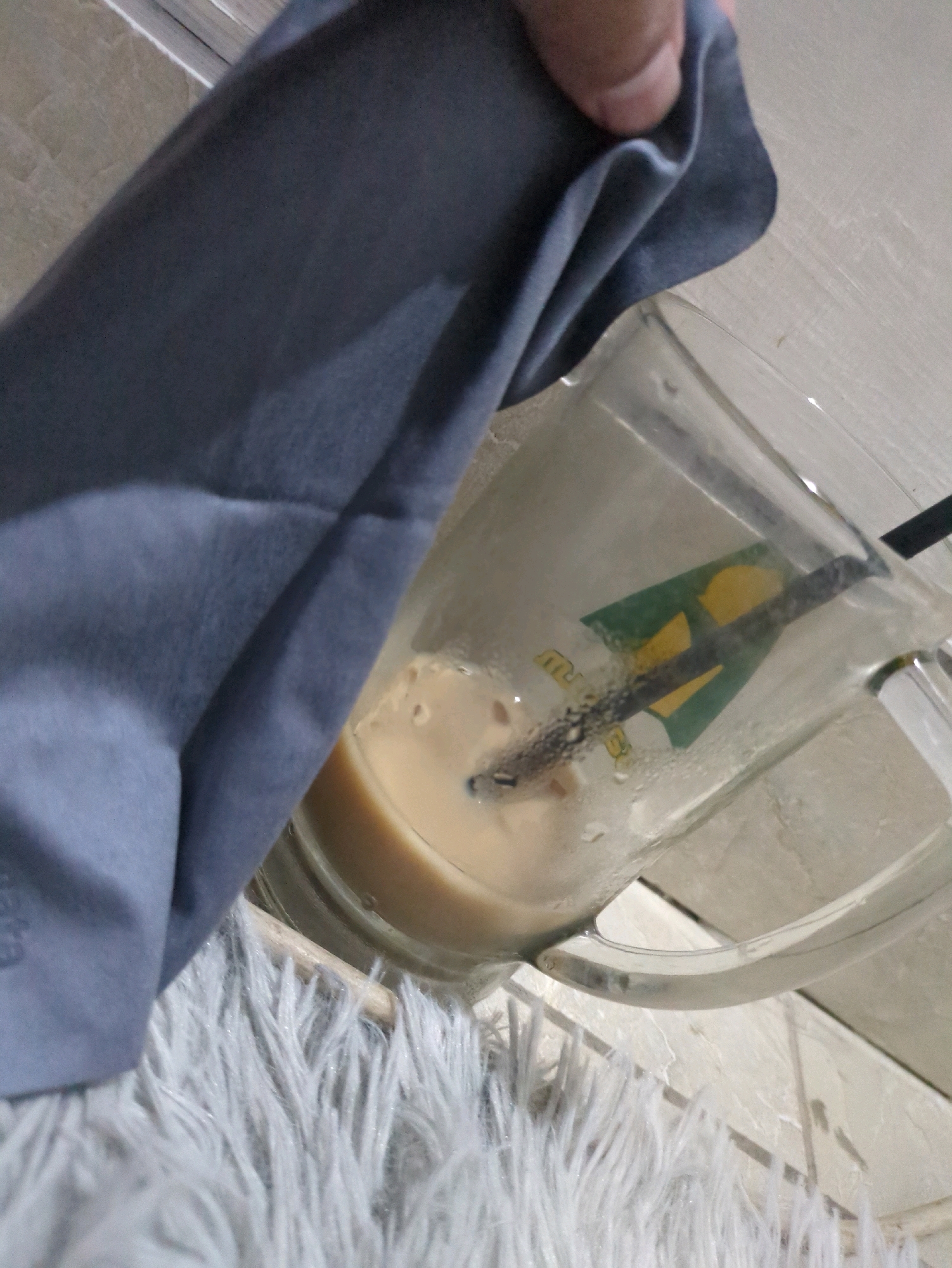 A close-up shot shows a glass mug filled with a light-colored liquid, possibly coffee or milk tea, with a black straw submerged in it. The mug has a logo on it with green and yellow colors. A hand is holding a gray cloth, partially obscuring the mug, as if wiping it. The background appears to be a tiled surface and a fluffy white rug. The scene is set in Sindon, Indonesia.