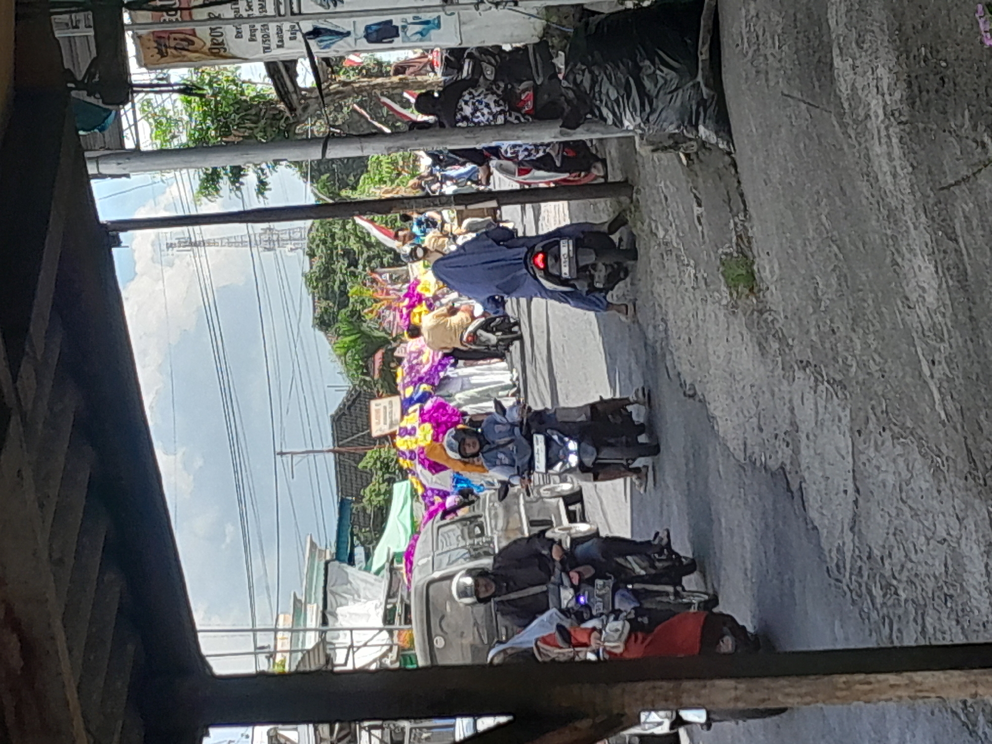 The image depicts a street scene in Gentengwetan, Indonesia, characterized by a procession of motorcycles and pedestrians. A dark-colored van is visible in the background, alongside a building with a billboard. The sky is overcast with clouds, and a cell tower can be seen in the distance. Several individuals on motorcycles are present, some wearing helmets and others without. One person is wearing a blue headscarf and riding a motorcycle. In the foreground, a large black plastic bag is placed on the ground. There are also colorful banners or decorations scattered throughout the scene, suggesting a possible event or celebration. The road surface appears to be asphalt.