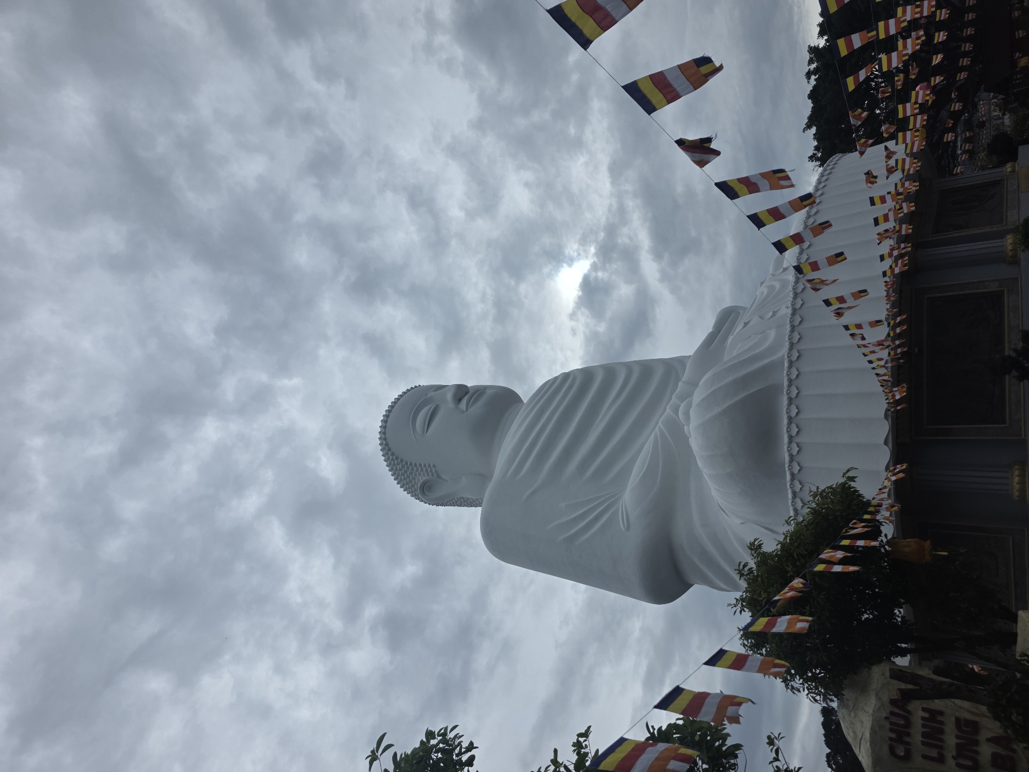 The image shows a large, white, seated Buddha statue. The statue is made of smooth, white material and is ornately decorated at its base. A string of colorful prayer flags, predominantly yellow, red, and white, is draped across the foreground and sides of the statue. The flags are attached to wires and hang in a descending pattern, with some fluttering in the breeze. Behind the statue, the sky is overcast with dramatic, swirling grey clouds. The bottom left of the image features a large, grey rock with text etched into it, and some trees and foliage. The text on the rock appears to be "CHÙA LINH ỨNG BÁ". This is the Linh Ung Pagoda, located in Da Nang, Vietnam.