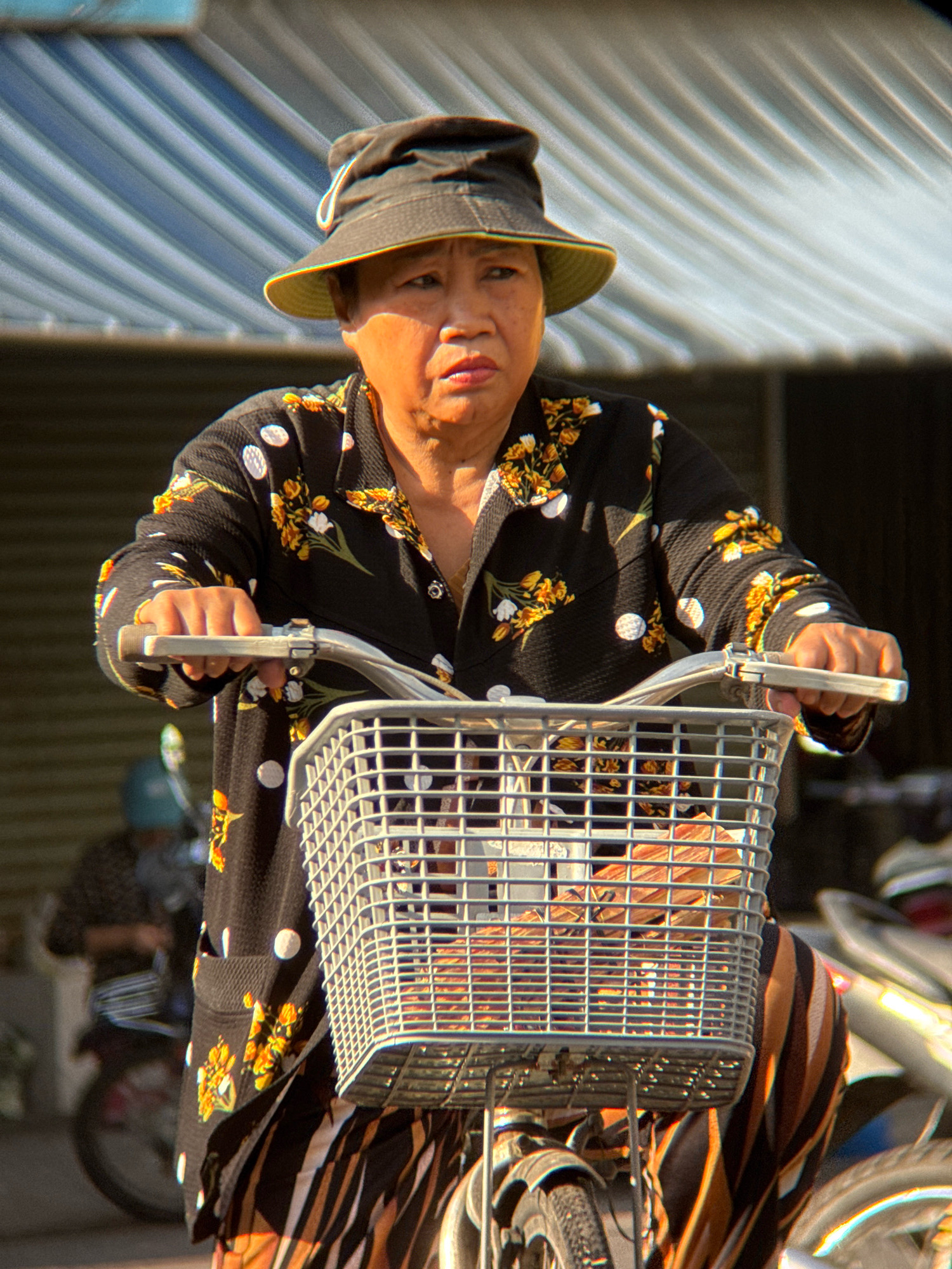A medium shot shows an East Asian woman wearing a wide-brimmed hat and a black shirt with a floral pattern. She is looking forward with a serious expression while riding a bicycle. The bicycle has a wire basket in front, containing some items. The woman's lower body is clad in a striped skirt. The background appears to be a street scene in Nha Trang, Vietnam, with a building structure featuring corrugated metal roofing. Other bicycles are visible in the background. The lighting suggests it is daytime.