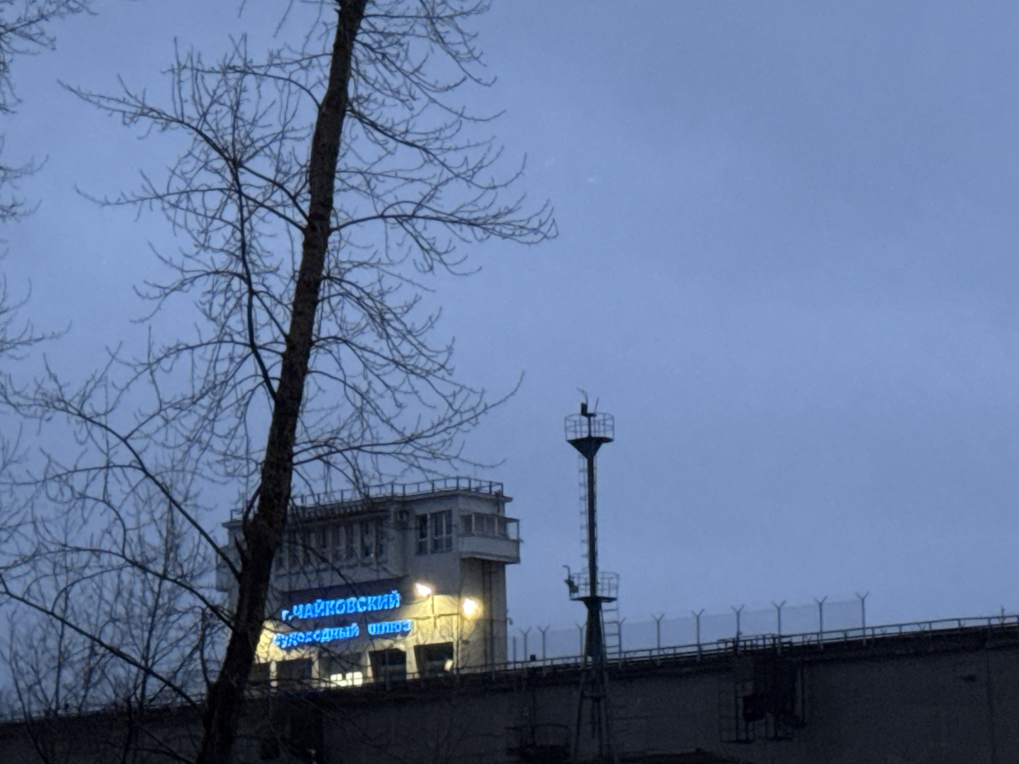 The image shows the Chaikovsky lock system on the Kama River in the city of Chaikovsky, Russia, at dusk. A bare tree stands in the foreground on the left. Behind the tree is a tall structure, with a control tower and blue Cyrillic lettering illuminated, presumably the name "Chaikovsky lock." A security fence with barbed wire stretches along the top of a concrete structure on which the lock is built, with tall metal security towers. The sky is overcast.
