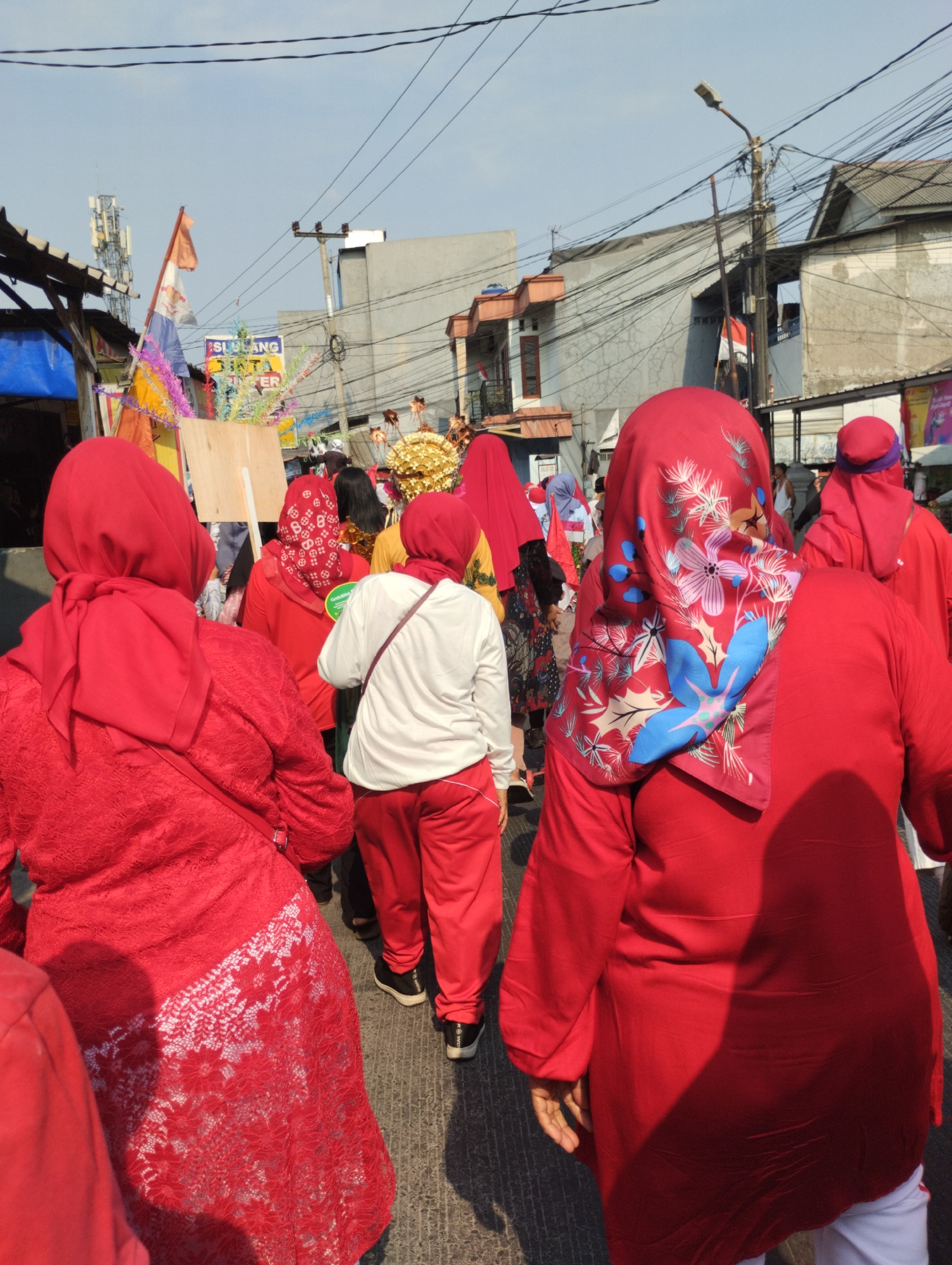 In Bekasi, Indonesia, a group of people, predominantly women, are walking in a procession on a street. They are all wearing red attire, with many also wearing red hijabs. Some of the hijabs have floral patterns. One woman in the foreground has a red lace overlay on her dress. In the background, there are buildings, power lines, and streetlights. There are also flags and colorful decorations, possibly part of a celebration or event. The sky is clear and blue. The scene appears to be taking place during the daytime.