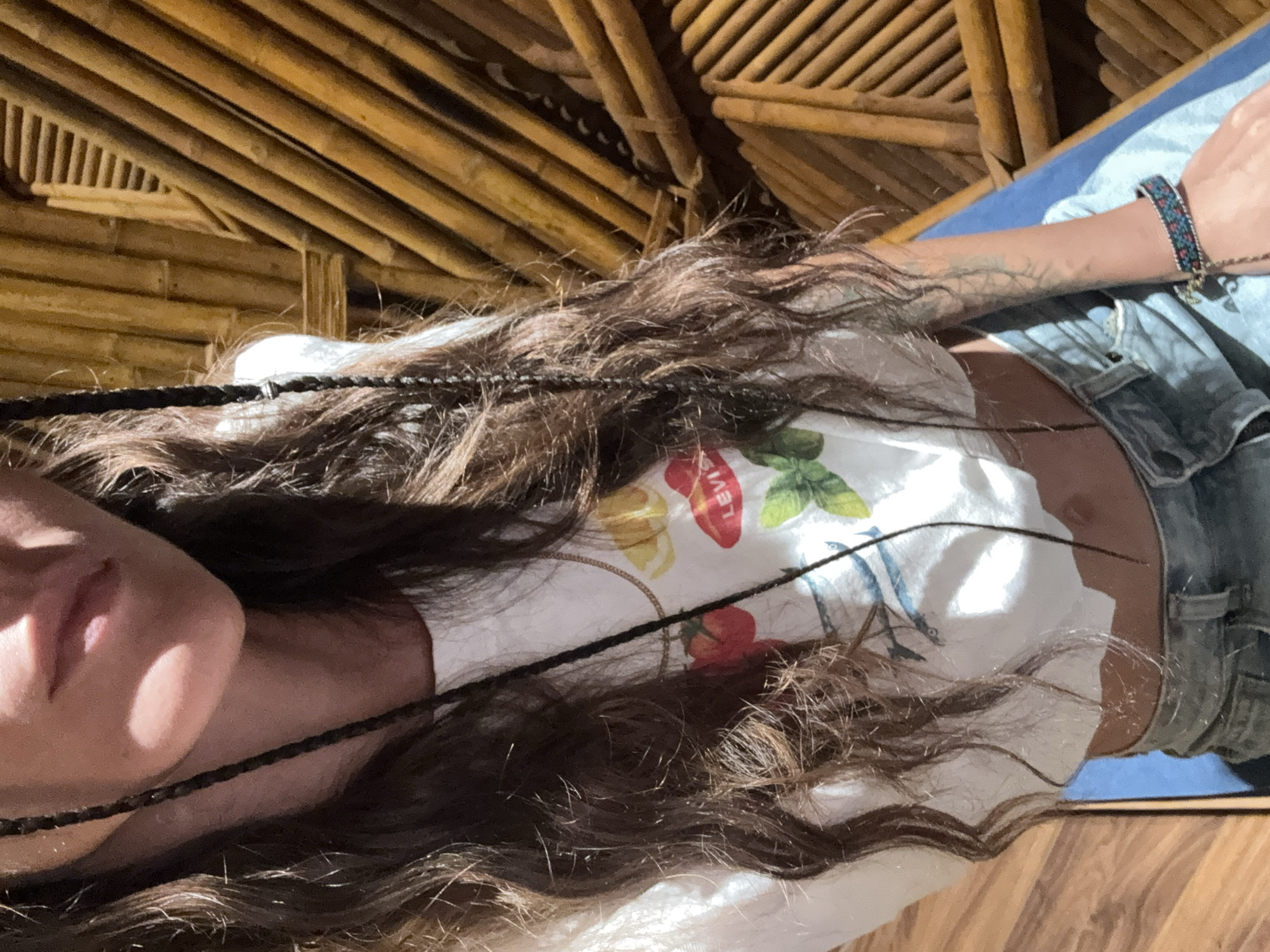 A close-up, downward-angled shot captures an individual, possibly female, from the upper torso to the lower face. Long, wavy, dark brown hair, interwoven with several thin dark braids, cascades over the shoulders and chest. The lower portion of the face is visible, featuring light-toned skin, slightly parted lips, and a distinct shadow cast under the chin.
The individual wears a white, short-sleeved, cropped t-shirt with a colorful graphic print. The print includes a red tomato, a yellow lemon slice, red lips with "LEVI'S" written in white, green leaves (possibly basil), and blue fish (likely anchovies or sardines). Light-wash denim jeans are worn low on the waist, exposing a small portion of the abdomen and navel. A thin gold chain necklace is visible around the neck. On the left wrist (viewer's right), a multi-colored woven bracelet with small charms is worn. A faint, branch-like tattoo is present on the inner left forearm.
The background features a textured wall or partition constructed from numerous warm, reddish-brown wooden (likely bamboo) poles and slats, arranged diagonally and horizontally, creating an intricate pattern. The floor beneath is planked wood of a lighter brown hue, with a blue mat or cushion positioned under the subject. Strong, warm directional light illuminates the scene from the left, casting prominent shadows. The scene is identified as taking place in Mù Cang Chải, Vietnam.