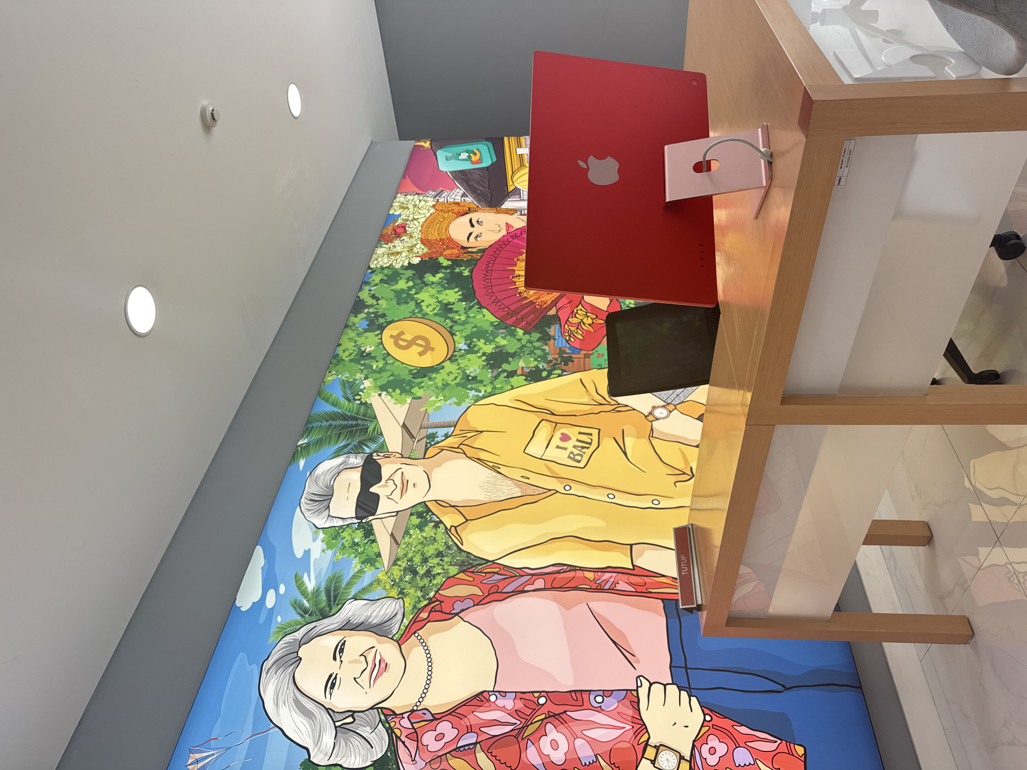 The image is taken inside a shop, potentially an Apple store, in Denpasar, Indonesia. A large colorful mural dominates the wall, featuring cartoon-like depictions of local people in traditional attire and scenes evoking Bali, like palm trees. A red iMac computer is positioned on a wooden desk in front of the mural. The mural includes depictions of older tourists wearing “I love Bali” shirts. There are other Apple products on the table. The store has a modern, minimalist aesthetic with a gray ceiling and recessed lighting. The floor has a light gray tile pattern.