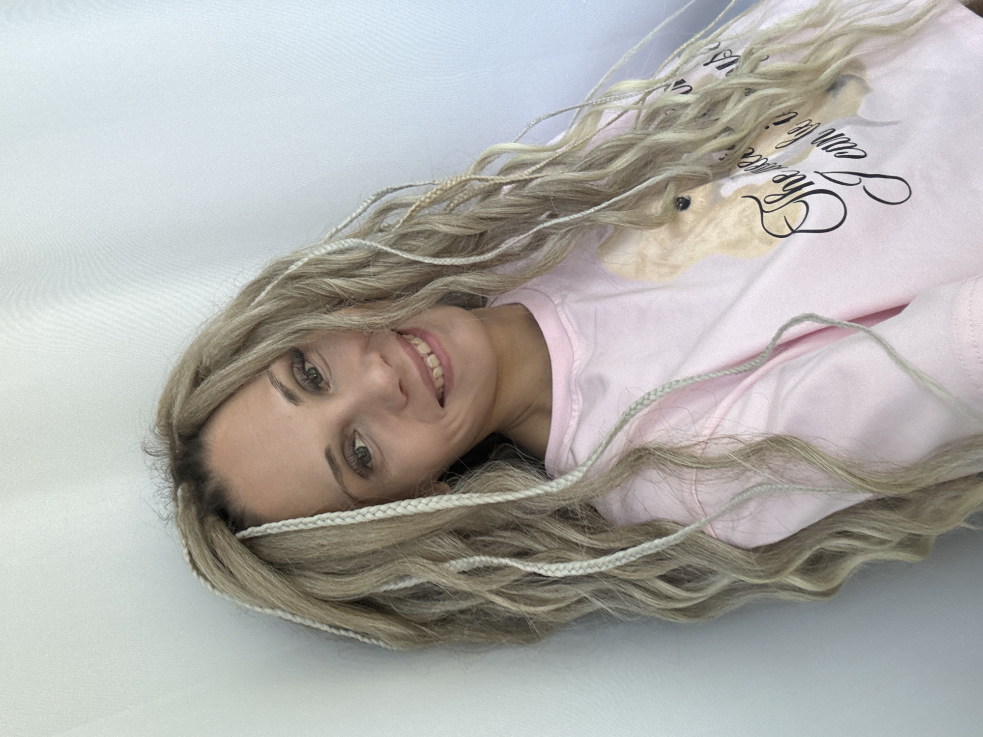 A woman with blond hair and braids is lying on a white surface. She is wearing a light pink sweatshirt with a graphic design on it. The woman is smiling and looking directly at the camera. The background is plain white. It is not possible to determine the location as Заволжье, Russia from this image alone.