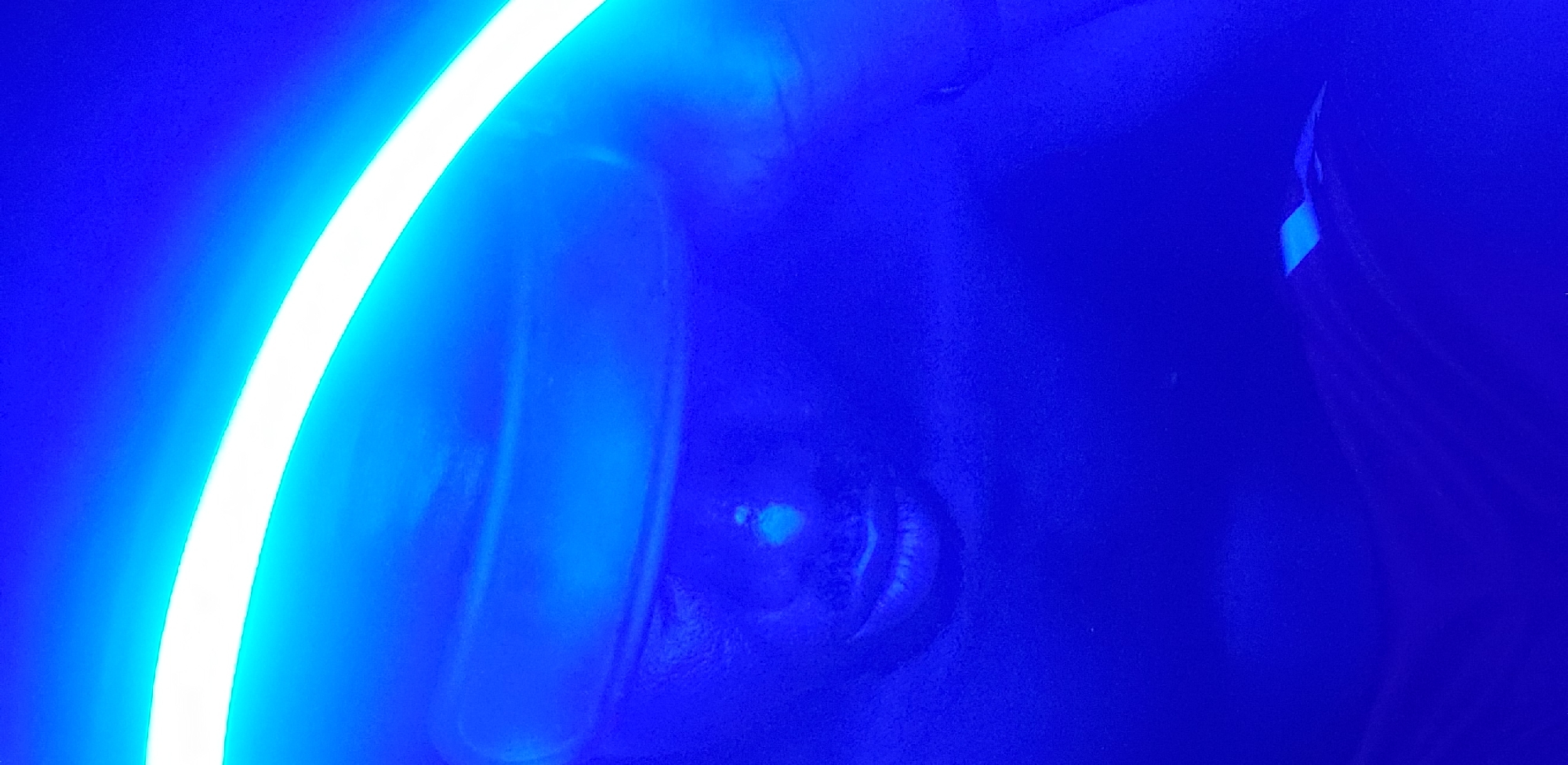 The image features a human face under intense blue lighting. A bright, curved neon light source is positioned near the left side of the frame. The face is tilted, with an eye visible, reflecting the light. The overall environment is dark, with minimal detail discernible besides the face and the light source. No specific details connect the image to Narayi, Nigeria.