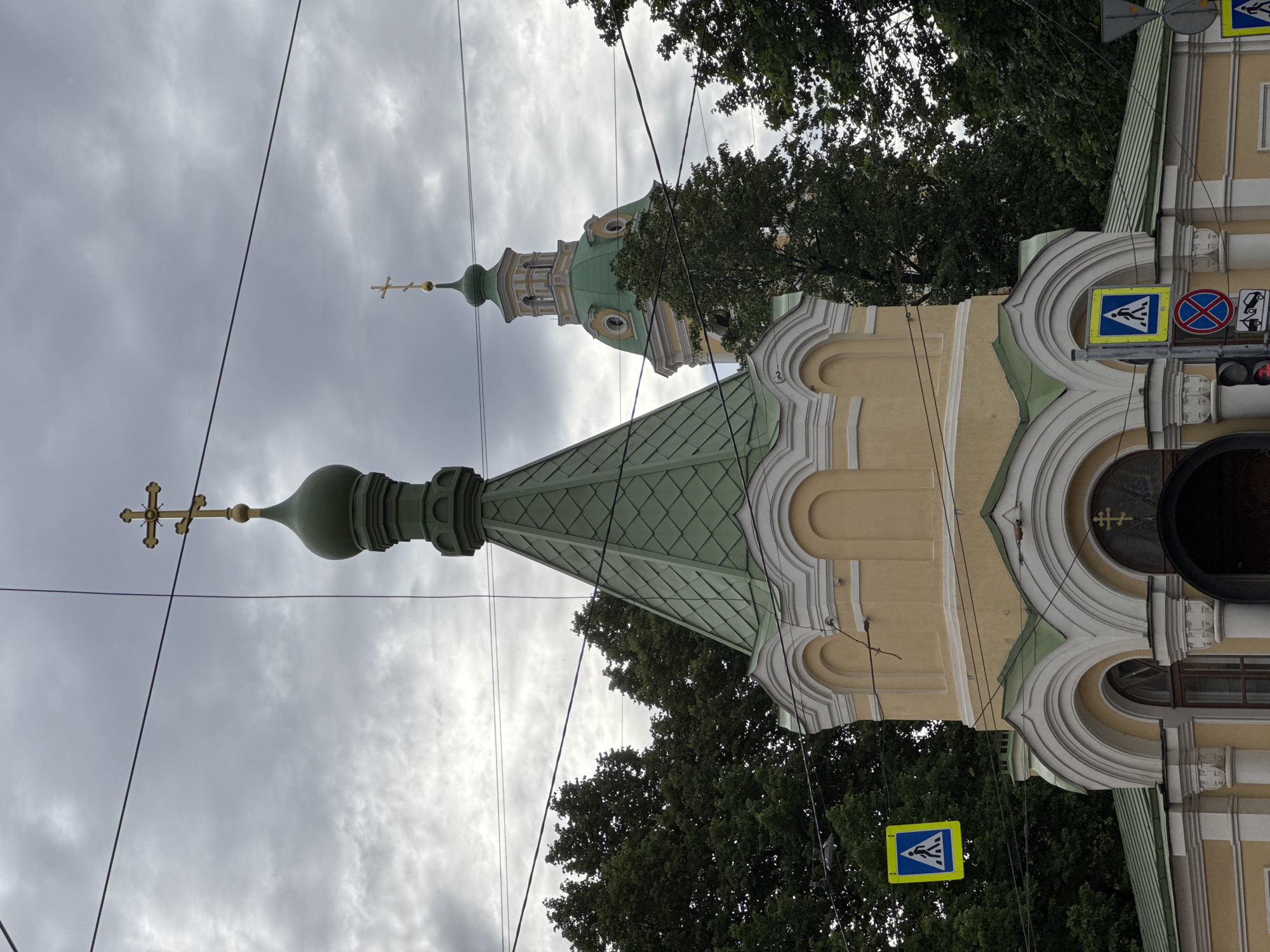 The image captures a view of the Smolny Cathedral in Saint Petersburg, Russia. The foreground features the onion dome of a smaller adjacent church or chapel, painted in green with a gold cross at its apex, contrasting with the pale yellow facade of the building. This structure is adorned with white decorative moldings around arched windows and doorways. Several pedestrian crossing signs, a common sight in urban environments, are visible, one on the left and another on the right side of the image. In the background, the distinctive green dome and bell tower of the Smolny Cathedral rise above a dense canopy of lush green trees. The sky is overcast, with a thick layer of grey clouds, suggesting a muted, possibly humid, atmosphere. Overhead, a network of black electrical wires cuts across the frame, adding a modern urban element to the scene. There are no people or animals visible in the image.
