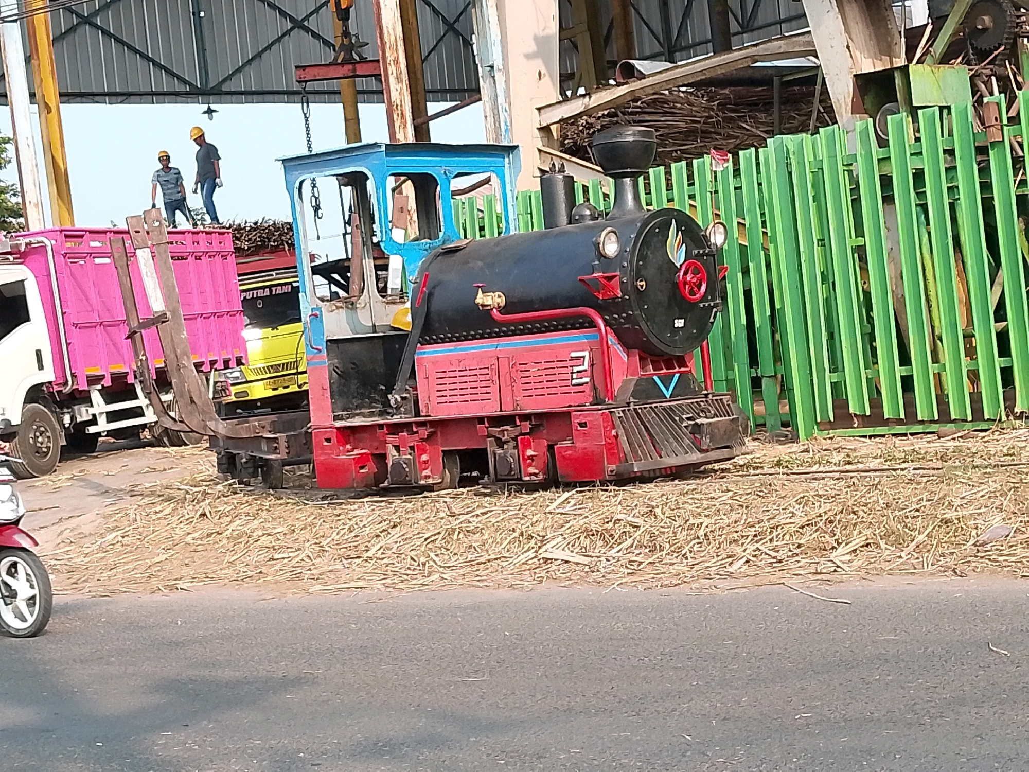 A miniature train with a black engine and red base, painted with red, blue, and yellow accents, sits on a pile of dry sugarcane stalks beside a bright green fence. To the left of the train is a pink truck and a yellow truck, both loaded with what appear to be bundles of sugarcane. Two individuals wearing yellow hard hats are standing on top of the pink truck, one holding a tool. A red motorcycle is partially visible in the lower left corner of the image. The scene is set in a clear, outdoor environment under a bright sky. The presence of sugarcane trucks and the train suggests an industrial or agricultural setting, possibly a processing facility. The location is identified as Geneng, Indonesia.