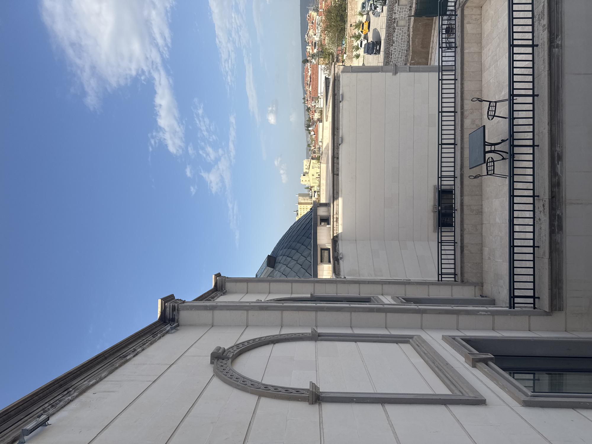 This is an exterior shot of a white stone building in Ürgüp, Turkey, with a balcony furnished with an iron table and two chairs. The building is multi-storied, featuring a domed roof and arched decorative elements. A fire escape ladder is attached to the building's side. In the background, the cityscape of Ürgüp is visible under a partially cloudy blue sky. Cars are parked on a street near the building. The photo is taken from a low angle, emphasizing the building's height.