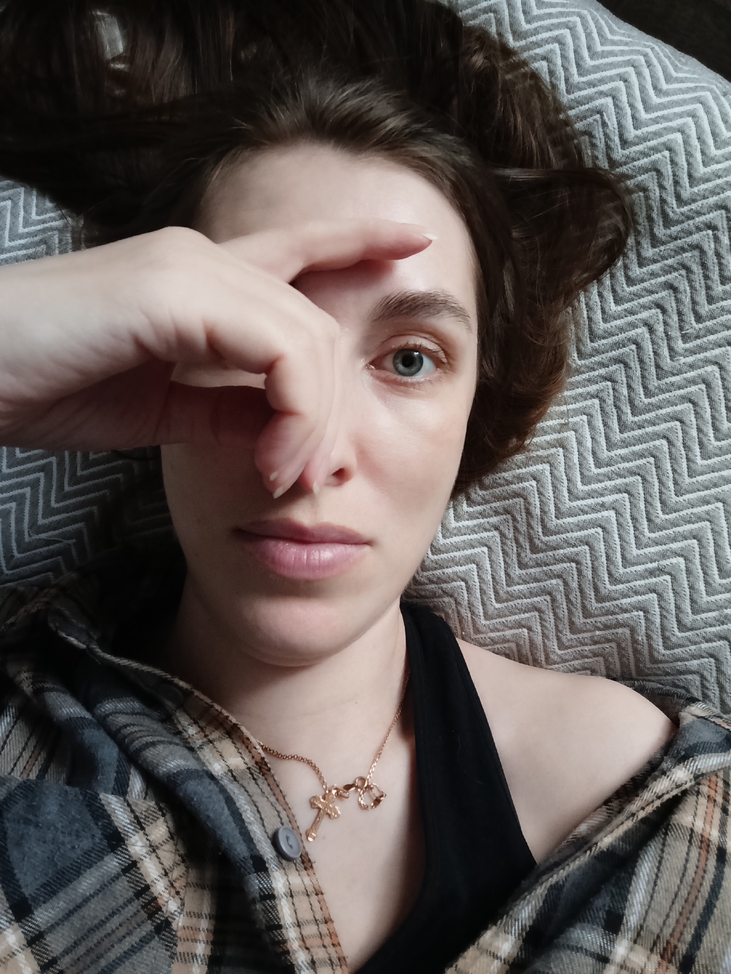 A fair-skinned person with brown hair is lying on a grey zigzag-patterned surface. They are wearing a black tank top under a grey, brown, and tan plaid shirt. A gold necklace with a cross pendant is visible. The person has their right hand raised, with fingers covering part of their face, obscuring one eye. Their visible eye is blue-grey. The image does not provide information about the location being Chaikovsky, Russia.