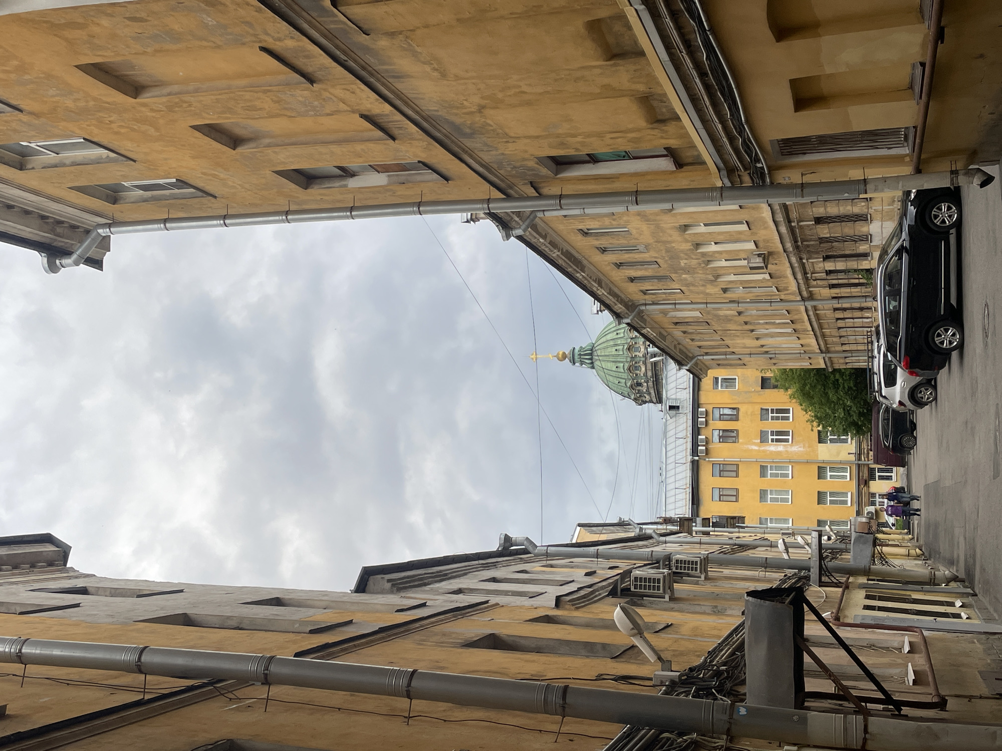 The image depicts a narrow alleyway between old, weathered yellow buildings in Saint Petersburg, Russia. A green dome with a golden cross atop it, belonging to St. Isaac's Cathedral, is visible in the distance, framed by the buildings. The sky is overcast with a gray, cloudy appearance. In the foreground, the textured walls of the buildings show signs of age and peeling paint, with various pipes and utility boxes attached to them. A few cars are parked in the alley. Two people are walking away from the camera in the middle ground. The overall scene conveys a sense of historical architecture and urban ambiance.