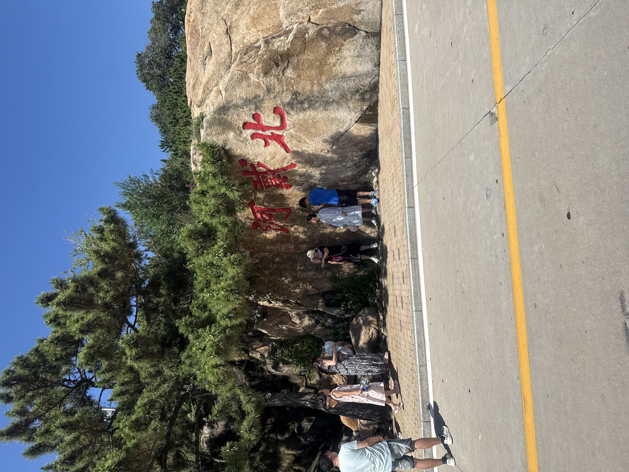 In the city of Qinhuangdao, China, a large rock face bearing red Chinese characters for "Hebei" is visible. Several individuals are gathered in front of the rock. On the left, a person wearing a light blue t-shirt and shorts stands facing away from the camera. To their right, a woman in a pink dress and another woman in a patterned dress are standing. A woman in a straw hat and a black dress is pointing to the right. Two young people, a boy in a blue t-shirt and shorts and a girl in a white dress, are standing near the center of the rock. The ground in front of the rock is paved with bricks, and a concrete road with a yellow line runs in the foreground. Lush green trees, including a large pine tree, overhang the scene. The sky is clear and blue.