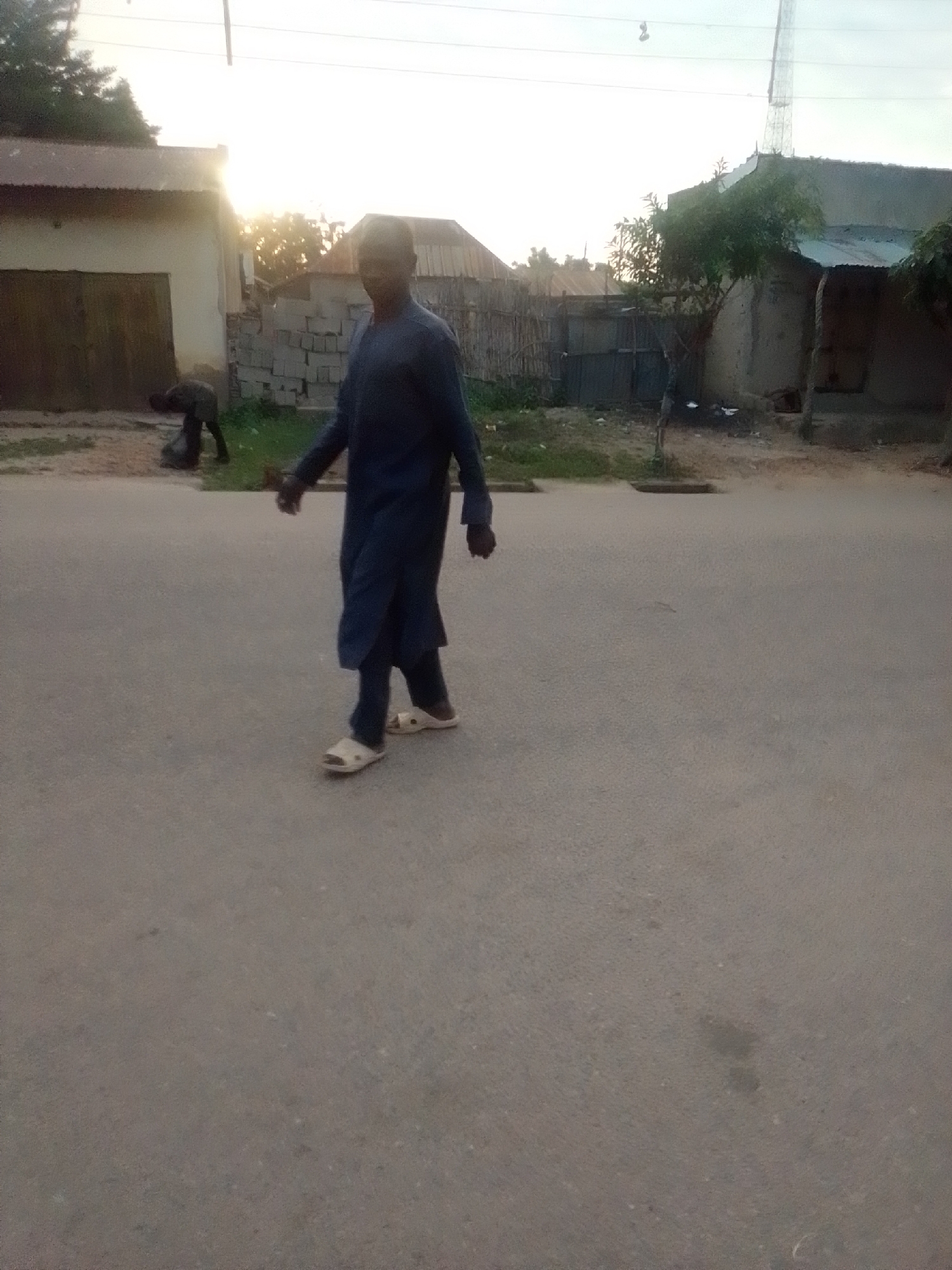 The image depicts an outdoor scene in what appears to be a developing urban or semi-urban area, potentially in Liji Hill, Nigeria. The lighting suggests it is either early morning or late afternoon, with the sun low in the sky, casting a warm glow. In the foreground, a man is walking across an asphalt road. He is wearing a dark blue, long tunic or traditional garment and light-colored sandals. His posture suggests he is moving forward, looking slightly to his left. In the background, to the left of the road, there is a building with a corrugated metal roof and large, dark doors. A second individual, partially obscured, is bent over near this building, appearing to be engaged in some form of manual labor, possibly involving a sack or debris. Further in the background, there are more structures with tin roofs, a fence made of corrugated metal or wood, and vegetation. A tall communication tower is visible in the far distance against the bright sky. The overall environment appears somewhat unkempt, with visible construction materials (blocks) and debris scattered around the periphery of the buildings.