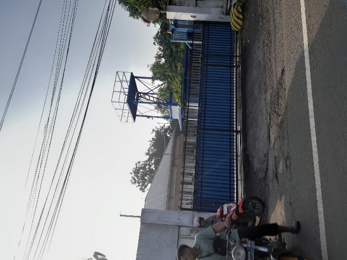 The image shows a view from the street in front of a gated building in Pungging, Indonesia (latitude: -7.497, longitude: 112.563). The building is closed off by a tall blue metal gate, and a large metal water tower stands to the left of the gate. There are numerous electrical wires running overhead, and a single white line extends from the lower right corner of the image towards the gate. Two men are standing on the street in front of the gate. One man is wearing a white shirt and black pants, and the other man is wearing a green shirt and black pants. The man in the green shirt appears to be holding a motorcycle. The building appears to be private, and there is a red and white flag hanging from the roof. The ground is uneven, and there are many potholes in the road. The sky is clear and blue.