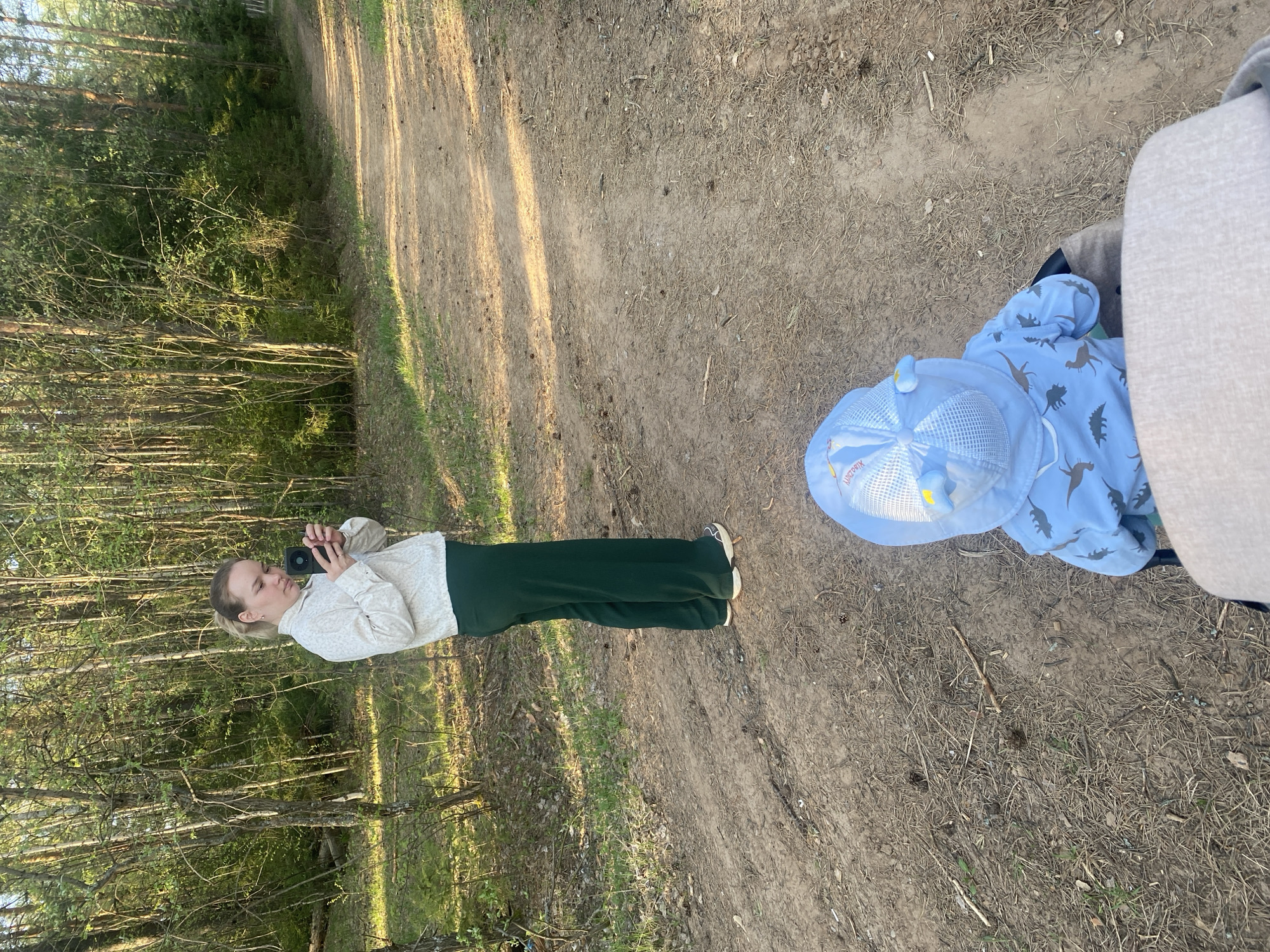 The image depicts a scene along a dirt path in a wooded area, likely in Марковский, Russia. On the left side of the path, a woman stands facing the forest and holding a smartphone in front of her as if taking a photo. She is wearing a light-colored, patterned top and dark green pants. To the right, a child wearing a blue dinosaur-patterned jacket and a matching hat sits in a stroller, with part of the stroller visible. The environment consists of a dirt path with foliage and trees on both sides, typical of a forest or park. The lighting suggests it is daytime.