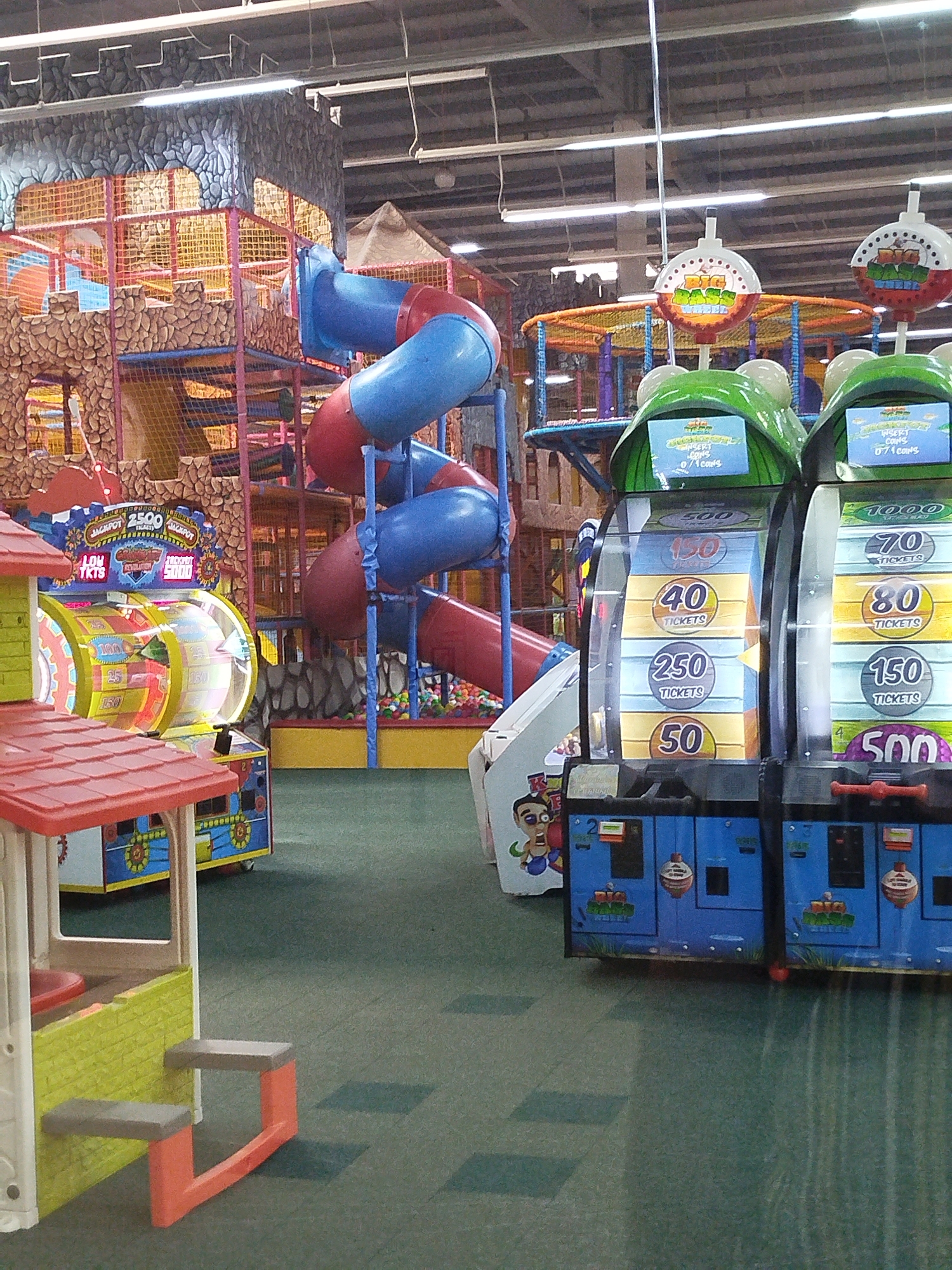 This is an interior photograph taken at a children's play center in Kremenchuk, Ukraine. The scene depicts an indoor playground structure featuring colorful slides and climbing areas. To the right of the playground, several arcade-style redemption games are visible. These games have screens displaying ticket amounts and slots for inserting coins. The flooring is a dark green carpet, and the ceiling has exposed beams and lighting fixtures. The overall environment appears to be a brightly lit entertainment venue designed for children. No individuals are clearly visible in the frame.