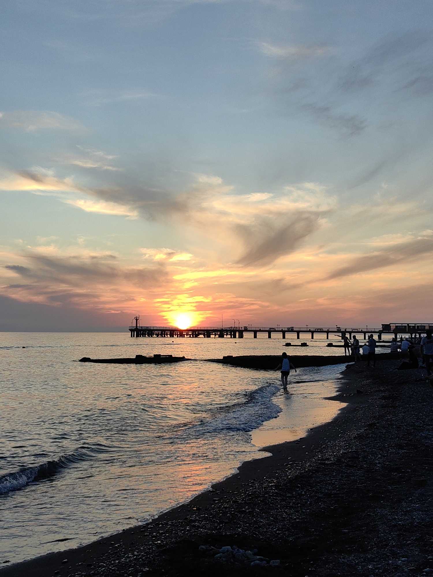The image displays a sunset scene off the coast of Sochi, Russia. A prominent orange-yellow sun is setting centrally, positioned behind a long pier structure that extends from the right into the sea. The sky transitions from light blue at the top to vibrant orange, pink, and yellow hues around the sun, with scattered clouds illuminated by the setting light. The calm sea reflects these warm tones towards the horizon and along the shoreline.
The pier is rendered in dark silhouette, supported by multiple columns, and includes railings and lights along its length. A larger structure or building is visible on the pier near the sun's position. The immediate foreground features a dark, pebbly beach on the right. A solitary human silhouette, appearing to be a child or small adult in a light top, walks into the shallow water on the left, facing away from the viewer. To the right, a group of several human silhouettes congregates on the beach and near the pier. Additionally, dark, low-lying structures are present in the water closer to the shore, and a small, dark floating object is visible further out to the left.