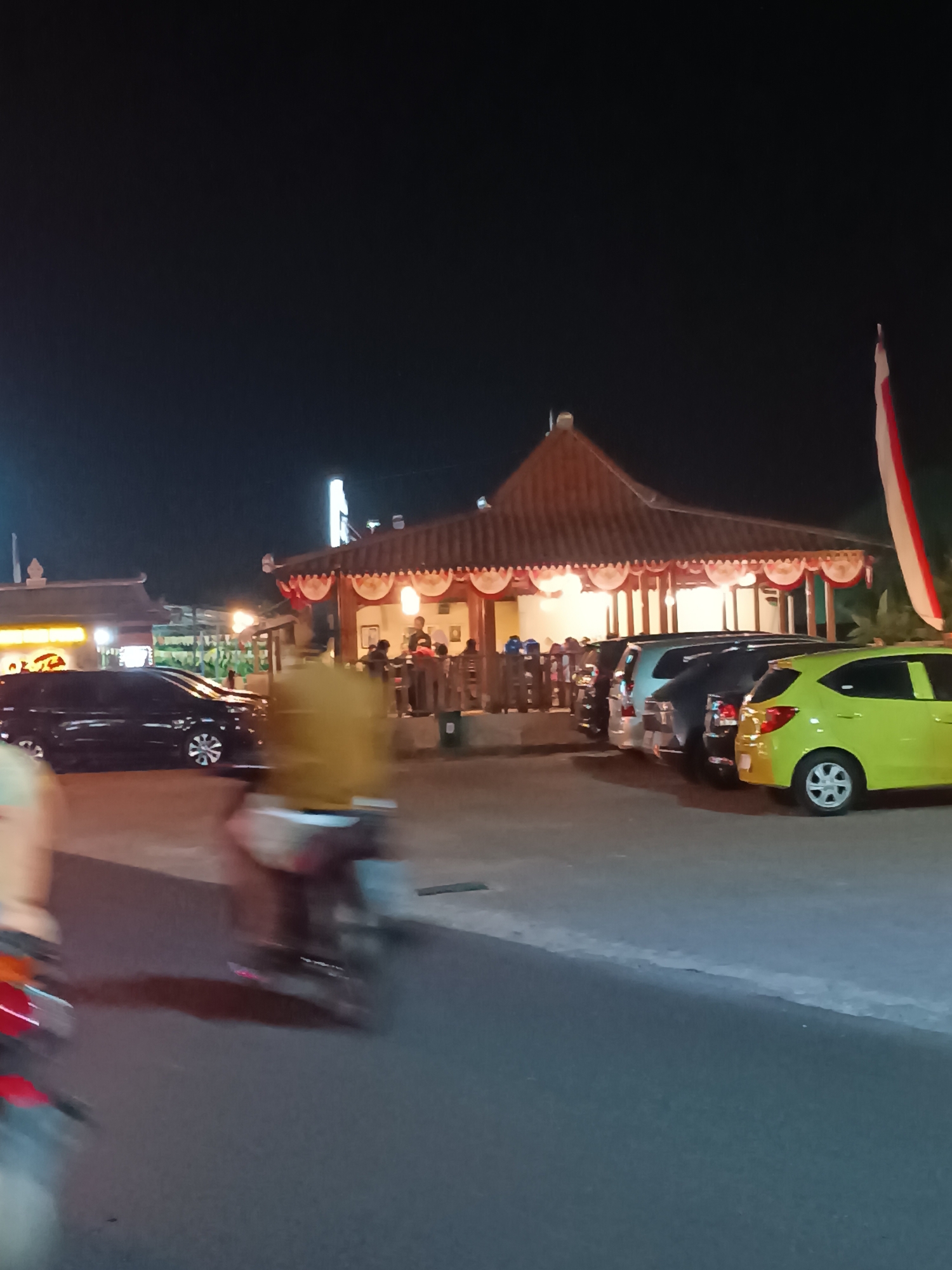 At night in Pleret, Indonesia, a scene unfolds with a prominent building featuring a tiered, angled roof adorned with red and white decorative scallops. Underneath this overhang, several people are seated at tables, illuminated by warm lighting emanating from within. A red and white flag, likely of Indonesia, flies from a pole to the right of the building. In front of the building, a row of cars is parked, including a bright yellow car on the right, a black SUV-like vehicle, and a lighter-colored SUV. To the left of the building, another dark car is parked. In the foreground, a motorcycle, partially blurred due to motion, is visible on the asphalt road, with a person seated on it. The environment appears to be an outdoor dining or gathering area, possibly a restaurant or warung, set against a dark night sky.