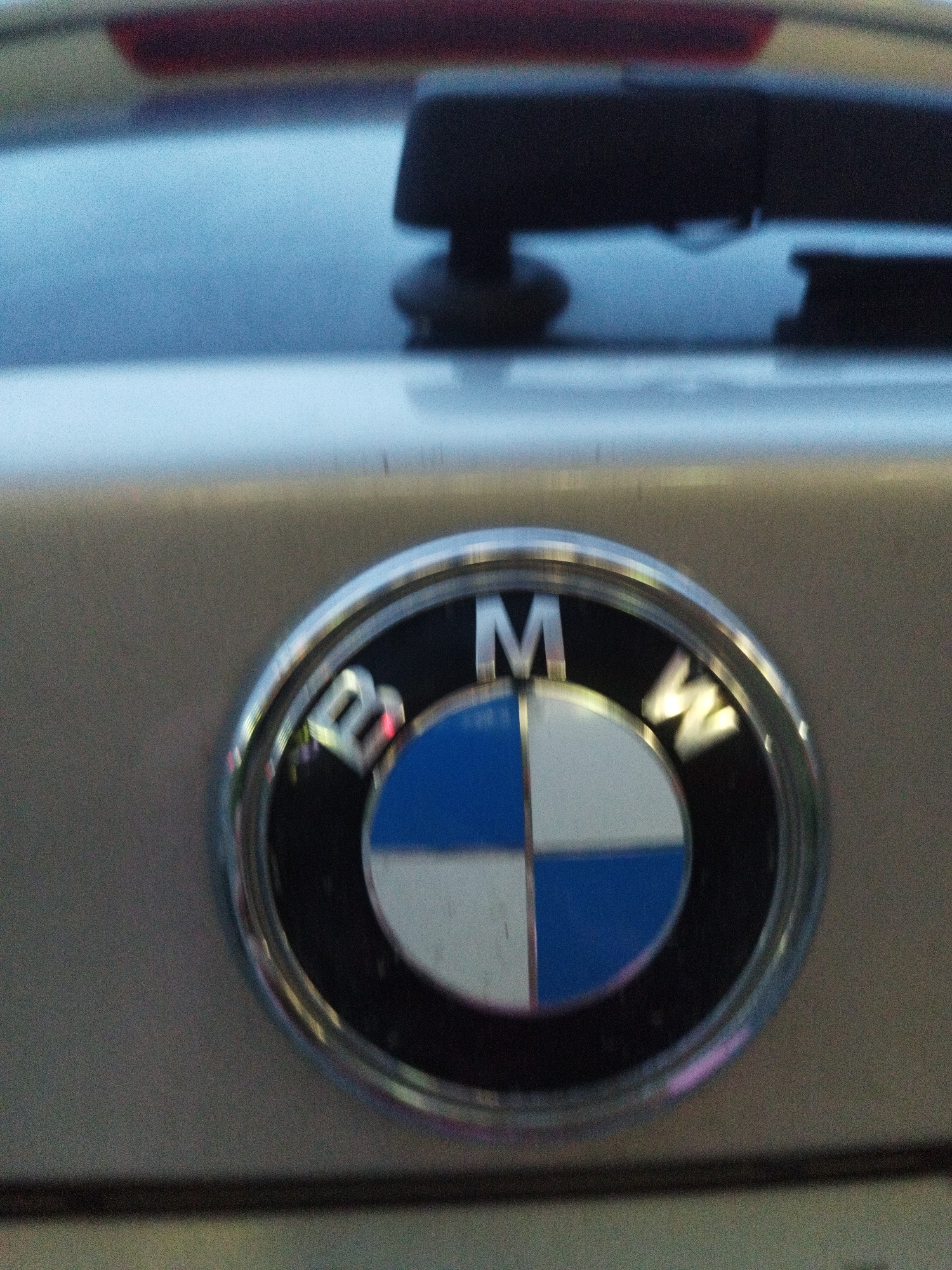 The image is a close-up, slightly blurry, medium-shot of a BMW emblem on the rear of a car. The emblem is circular with a chrome outer ring and a black inner ring. Inside the black ring are four quadrants: two blue and two white. The letters "BMW" are visible in white above the quadrants. The car's body appears to be a light metallic color. In the background, out of focus, is the rear window of the car, which is dark and reflects a red object, possibly a brake light. There is also a black object on the rear window, which might be a spoiler or a wiper. The lighting suggests it is daytime, possibly overcast or dusk, given the diffused light and faint reflections. No specific details about the city of Surgut, Russia, are discernible from the image itself.