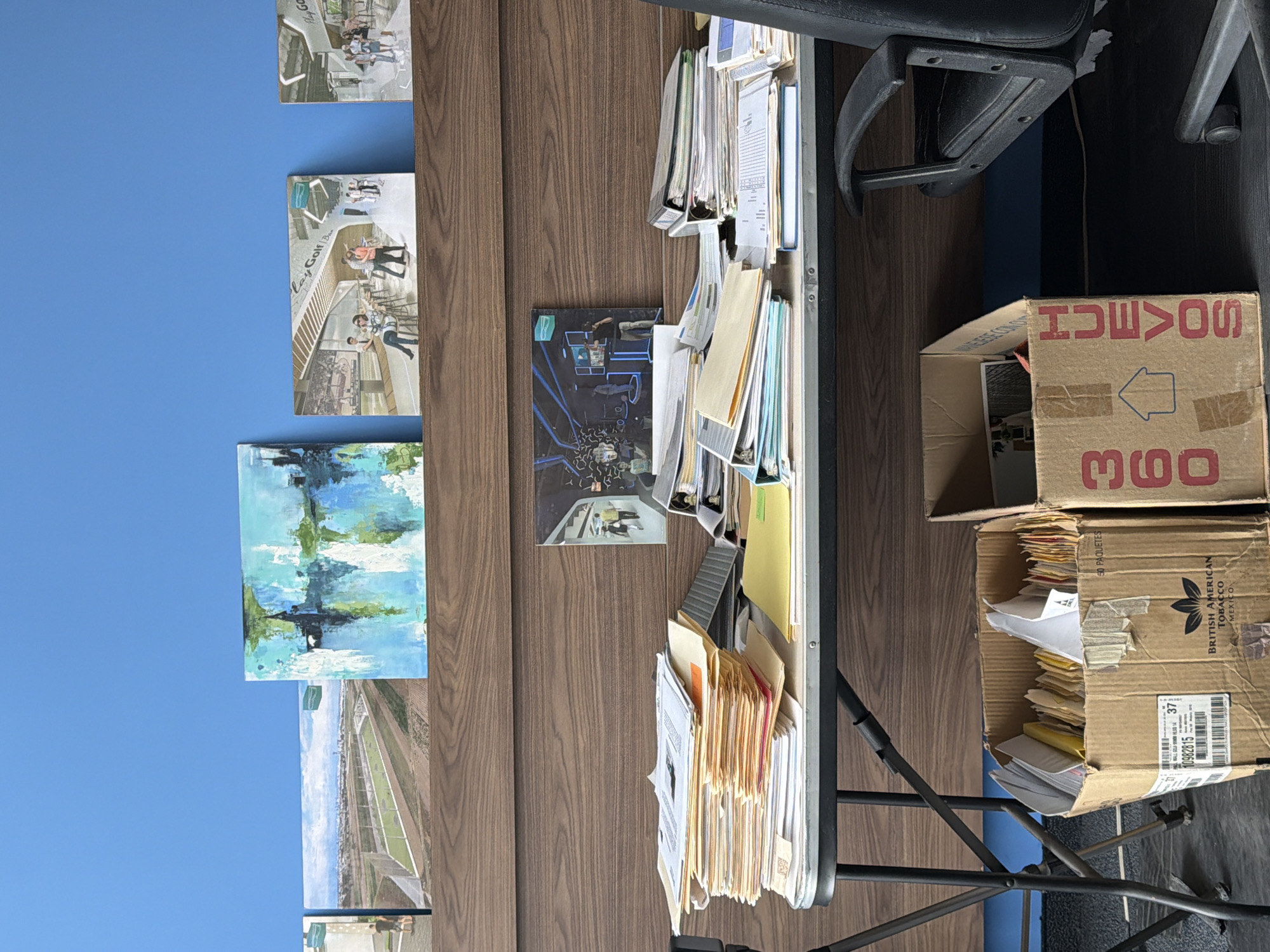 The image shows a cluttered desk area against a brown wood-paneled wall. Several framed artworks adorn the wall. The desk is laden with stacks of papers, files, and binders. Next to the desk, two open cardboard boxes are filled with documents and envelopes. One box is labeled "HUEVOS 360" and the other features a label with "BRITISH AMERICAN TOBACCO MEXICO" along with the text "50 PAQUETES", and a barcode with "10982815 37". The labels "Lay Golf Bar" and "Flay Go" are visible on two of the frames. An office chair is also visible near the desk. The location, San Bernardino Tlaxcalancingo, Mexico, is not explicitly evident in the image, but the presence of the British American Tobacco box suggests a possible connection to the location.
