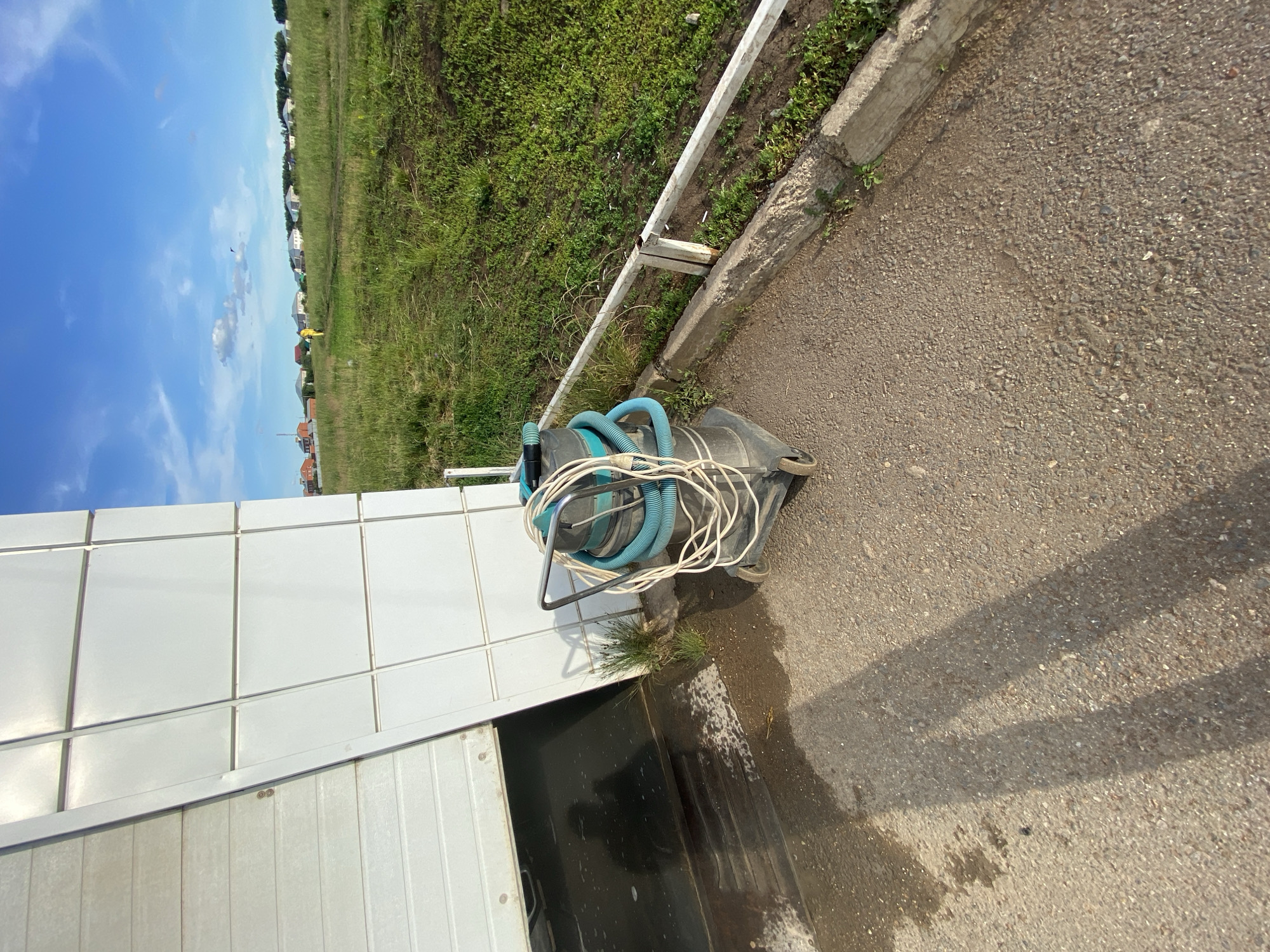 The image depicts an outdoor scene in the city of Dyurtyuli, Russia, likely on a sunny day with a clear blue sky and scattered white clouds. In the foreground, a grey industrial vacuum cleaner with blue hoses and white power cords wrapped around it rests on a paved surface. The vacuum cleaner is on wheels and has a metal handle. Adjacent to the vacuum cleaner is a large, white, sectional garage door, partially open, revealing a dark, wet interior. The ground in front of the garage door is wet and appears to be concrete with some patches of dirt and grass. To the right of the vacuum cleaner, a low white metal fence runs along a concrete curb separating the paved area from a grassy field. The field is lush and green, with some dirt paths winding through it. In the middle ground, in the distance, a person wearing a bright yellow jacket is visible walking across the grassy field. Further in the background, a row of houses with red and grey roofs lines the horizon, suggesting a residential area or the outskirts of a town. The overall environment suggests a utilitarian or industrial setting adjacent to a more natural, open space.
