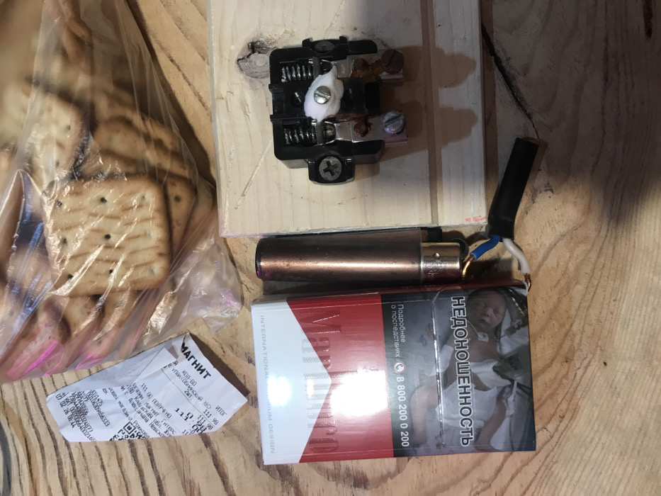 The image displays a collection of personal items laid out on a light-colored wooden surface, possibly a table or floorboards. The lighting suggests an indoor setting, and the overall impression is casual and somewhat utilitarian.
On the left side of the frame, there is a clear plastic bag filled with numerous square-shaped crackers or biscuits. Many of the crackers have small holes and a slightly toasted appearance. Below the bag, a crumpled white paper receipt is visible.
In the center of the image, a pack of Marlboro cigarettes is prominently displayed. The pack features the brand's distinctive red and white design, along with a graphic health warning on the visible side, depicting a premature baby. Above the cigarette pack rests a copper-colored metallic cylinder, resembling a large lighter or a battery housing. Exposed blue and white electrical wires are connected to one end of this cylinder, leading off to the right.
At the top center, an electrical component, possibly a switch or a relay, is mounted on a small, light-colored wooden block. This component is black, with visible springs, screws, and metal contacts that show some signs of rust or wear. A small piece of white, fluffy material (possibly cotton or insulation) is wedged near one of the screws.
Visible text from the items includes:
* **On the cigarette pack:** "НЕДОНОШЕННОСТЬ" (Prematurity), "Подробнее о последствиях" (More about the consequences), a phone number "8 800 200 0 200", and "INTERNATIONALMIUM DESIGN" (partially visible) along with the Marlboro brand logo.
* **On the receipt:** The name of the supermarket "МАГНИТ" (Magnit) is clearly visible at the top. Other details include "КОЛ-ВО" (Quantity), "В упак Обнинский хо", a price "111 00 ПОЛУЧЕНО" (111.00 RECEIVED), "газин Магнит ТитеPC", a location "180 Kalyская обл. и с. Александра Hesickor" (Kaluga region and Alexander Nesickor village), and a "итог 111.98" (Total 111.98). Various numerical codes and timestamps are also present.
The arrangement of items, particularly the exposed wires connected to the metallic cylinder and the electrical component, suggests a possible improvised electrical setup or a charging station among everyday consumables.