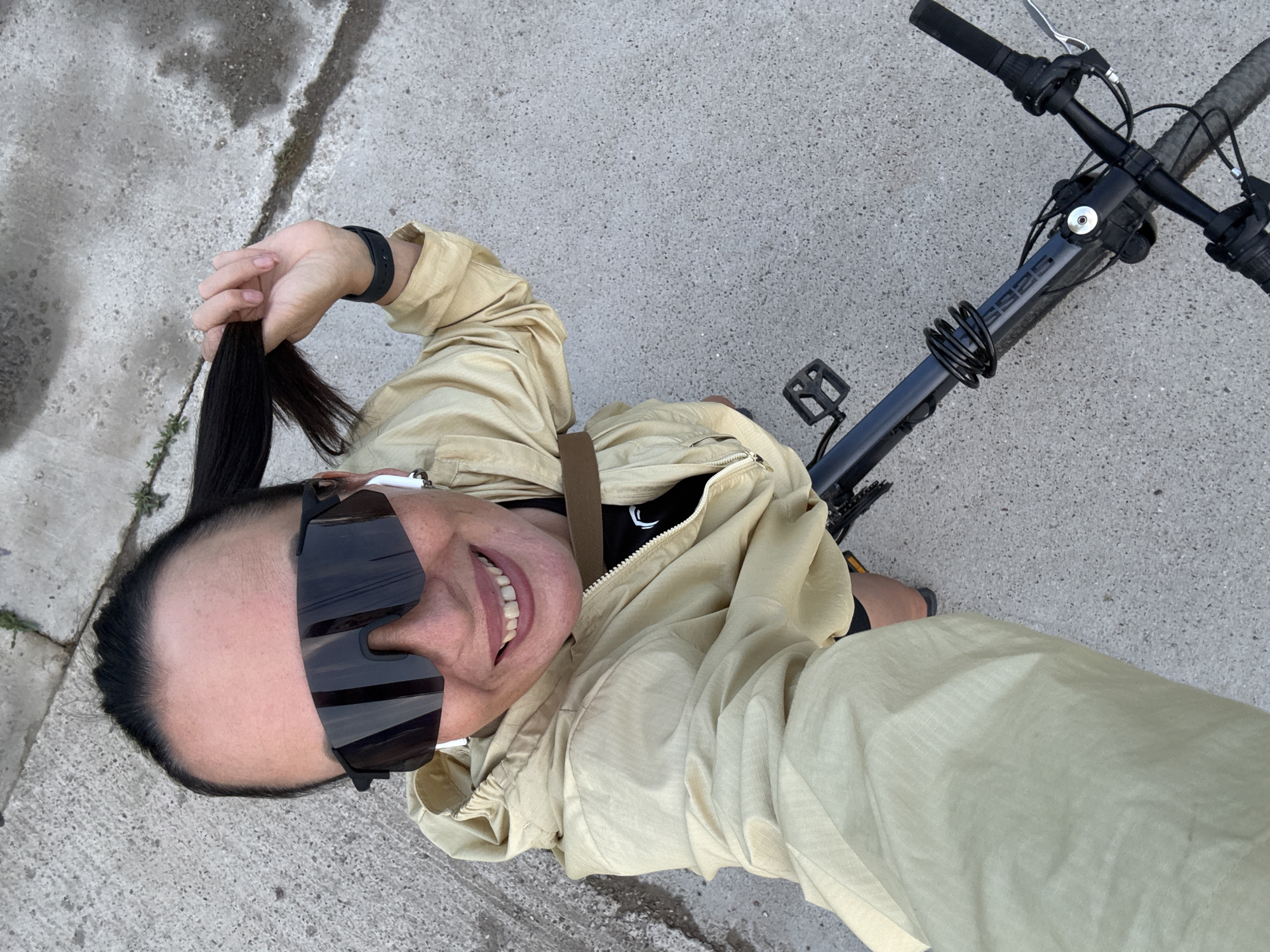 An overhead, selfie-style shot shows a woman from the chest up, standing outdoors on a paved surface, likely a sidewalk or pathway, with a bicycle positioned in front of her. She is wearing a tan, hooded jacket, black sunglasses, and white wireless earbuds. Her dark hair is pulled back, and she is holding a section of it in her right hand. The woman is smiling, with her teeth visible. A portion of the bicycle's handlebars and frame is visible in the lower part of the image. The environment appears to be urban or suburban, with concrete slabs visible on the ground. The image was taken in Chaikovsky, Russia.