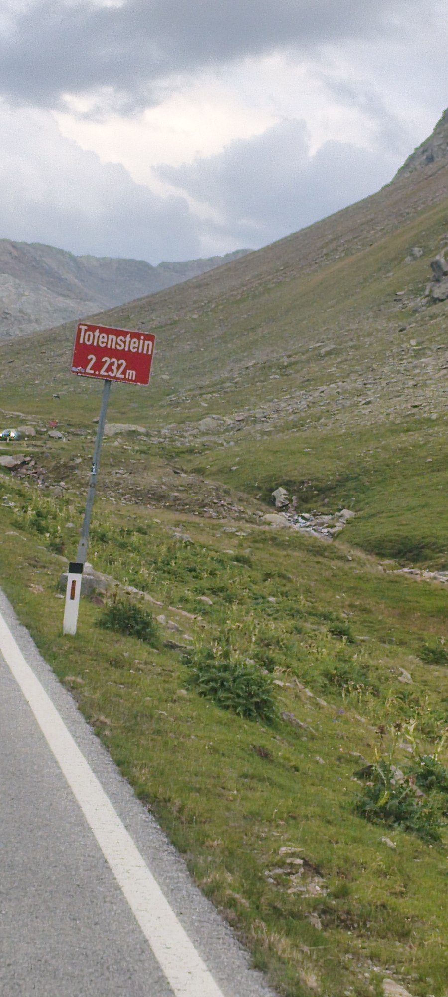 This is a vertical, medium-distance, eye-level shot of a mountainous landscape in Sölden, Austria. In the foreground, a paved road with a white line runs along the left edge of the frame. To the right of the road, a grassy, rocky slope rises steeply, dotted with sparse green vegetation. A signpost with a red rectangular sign reading "Totenstein 2.232m" stands prominently on the slope. The sign indicates the name "Totenstein" and an altitude of 2,232 meters. Further up the slope, the terrain becomes more rugged with larger rocks and scree. The background is dominated by the hazy shapes of distant mountains under a cloudy sky. The overall atmosphere is overcast and suggests a cool, high-altitude environment. There are no people visible in the frame.