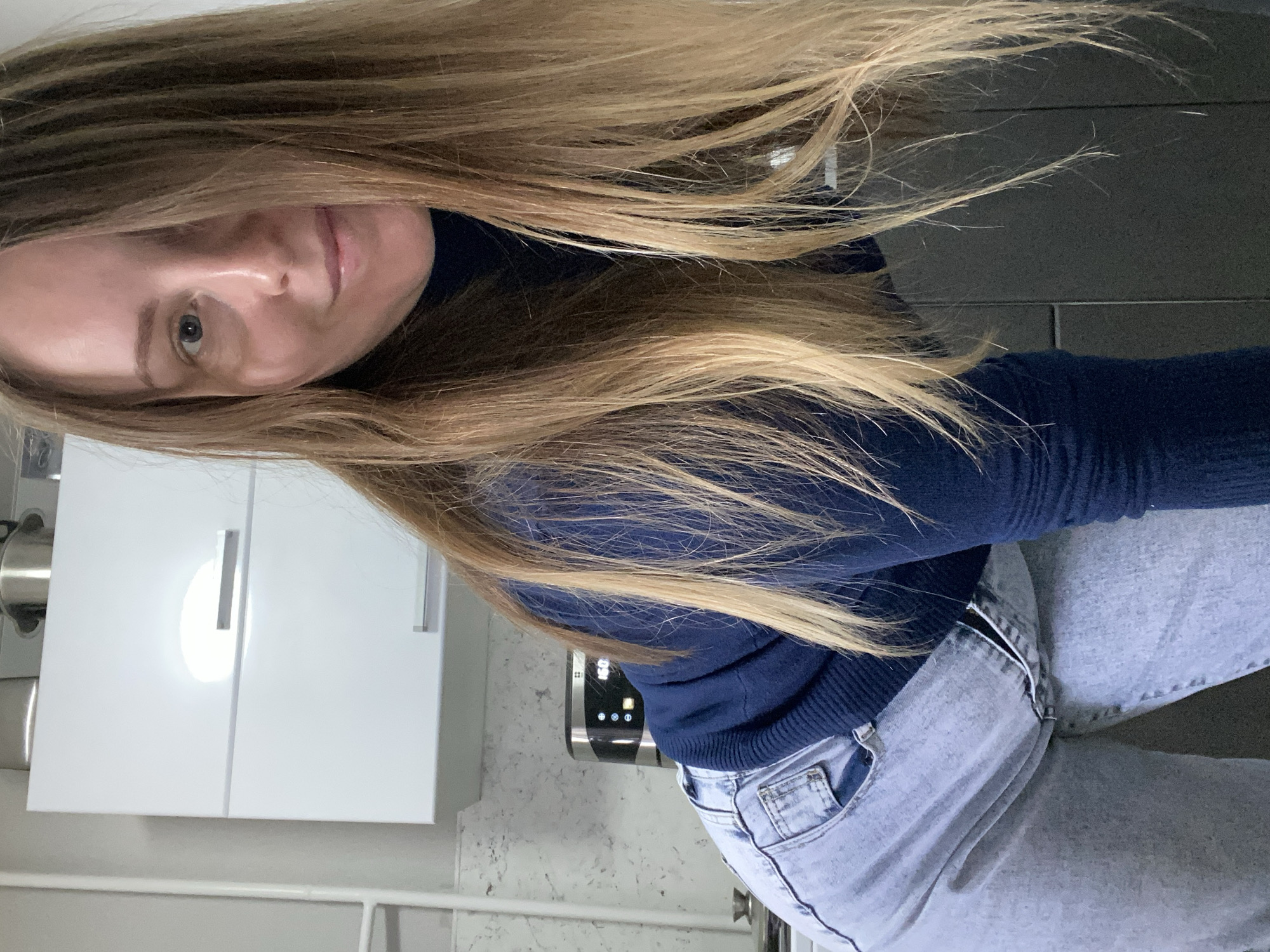 The image features a woman in an indoor setting, likely a kitchen, presented in a portrait orientation but rotated 90 degrees clockwise. Her long, flowing light brown hair, with lighter highlights, dominates a significant portion of the frame, cascading over her shoulder and face. She looks towards the camera with a neutral to slightly smiling expression, revealing part of her face, including one light-colored eye.
She is dressed in a navy blue, long-sleeved top and light-wash blue jeans. In the background, to the left, are modern white kitchen cabinets with integrated handles, one reflecting a bright light source. Behind her right arm, a silver and black appliance with a digital display and circular buttons is partially visible. The walls or countertops in the background feature a light, speckled pattern, suggesting a modern kitchen design. Another section of the wall appears darker grey with subtle horizontal lines, possibly indicating tiling. The scene is well-lit, but the specific time of day is not discernible. No legible text is visible in the image.