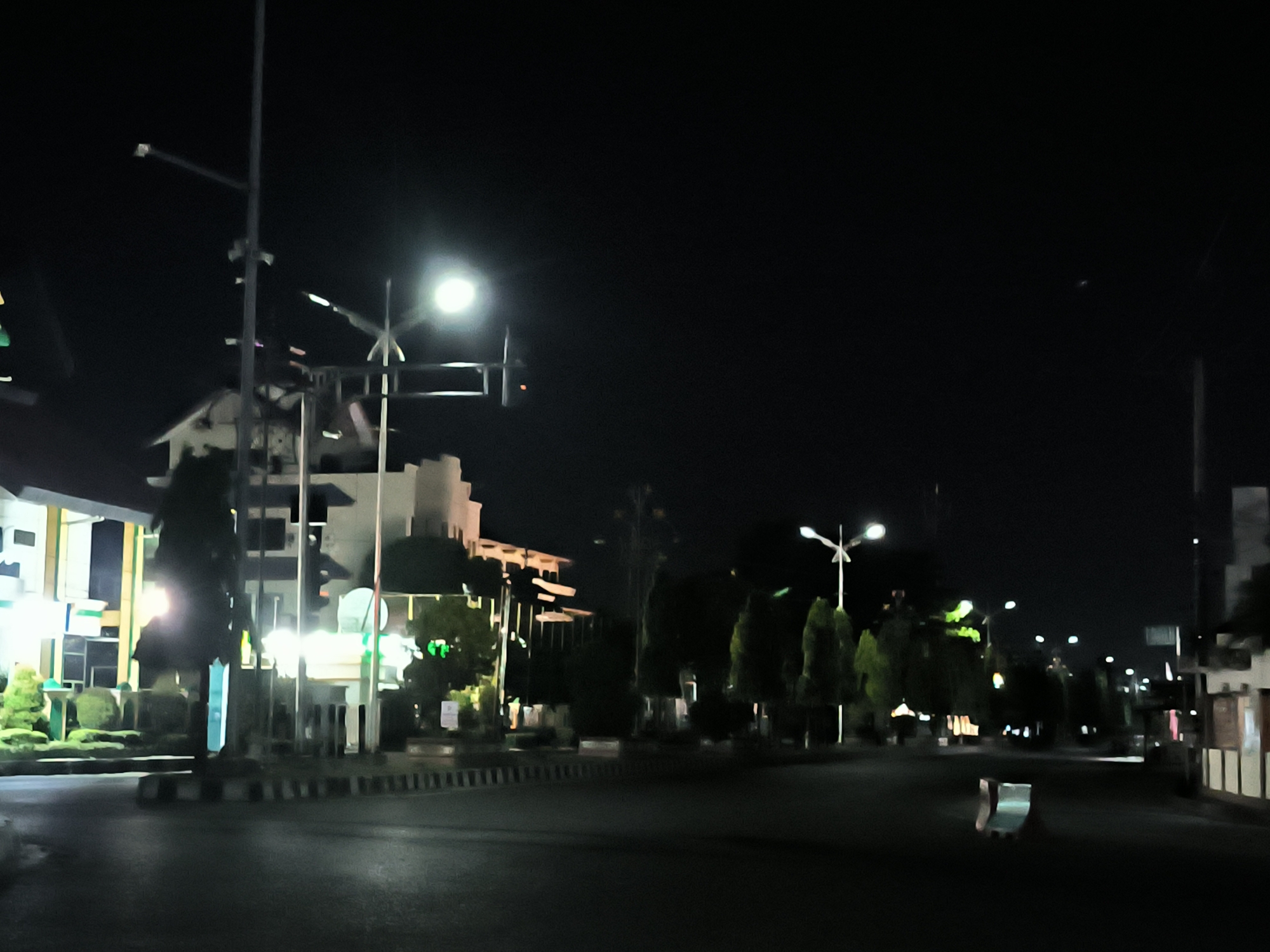 The image depicts a street scene at night in Lhokseumawe, Indonesia. The street is illuminated by streetlights. Buildings can be seen on the left side of the image. There are trees and other greenery lining the street. No people are visible. The road surface appears wet, possibly from recent rain.