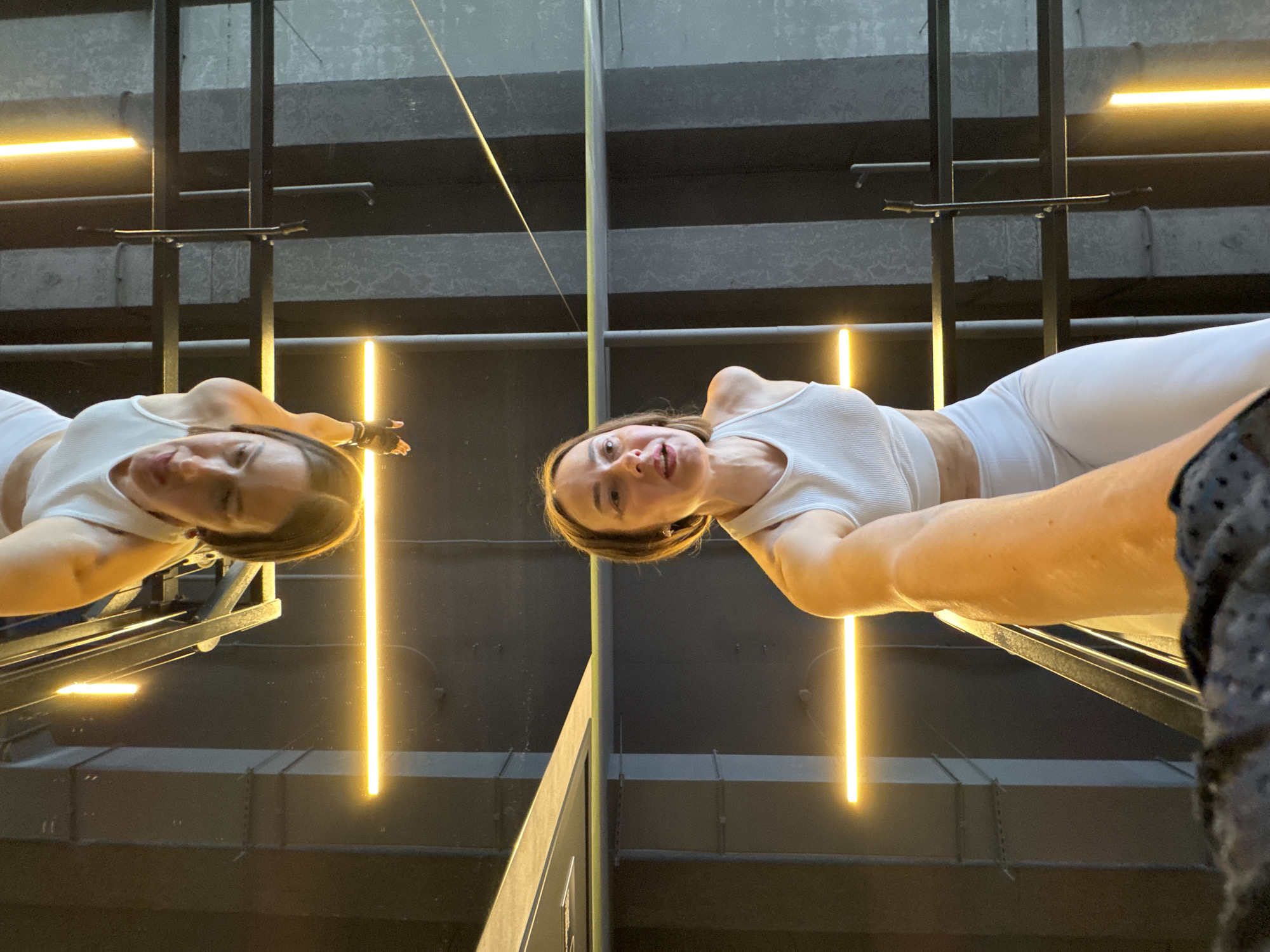 A woman is captured in a mirror selfie, her image reflected vertically. She is wearing white athletic wear, a ribbed tank top and leggings, and her hair is tied back. She is lying on a mirrored surface, looking up at the camera, with a slight smile. Her left arm is extended, and she is wearing a black glove on that hand. The background appears to be a gym or fitness studio, with industrial-style ceilings and vertical yellow strip lighting. The lighting creates a dynamic and modern atmosphere. The reflection shows a parallel structure of the gym equipment, adding depth to the image. The scene appears to be indoors, and the time of day is not discernible. No visible text is present in the image.