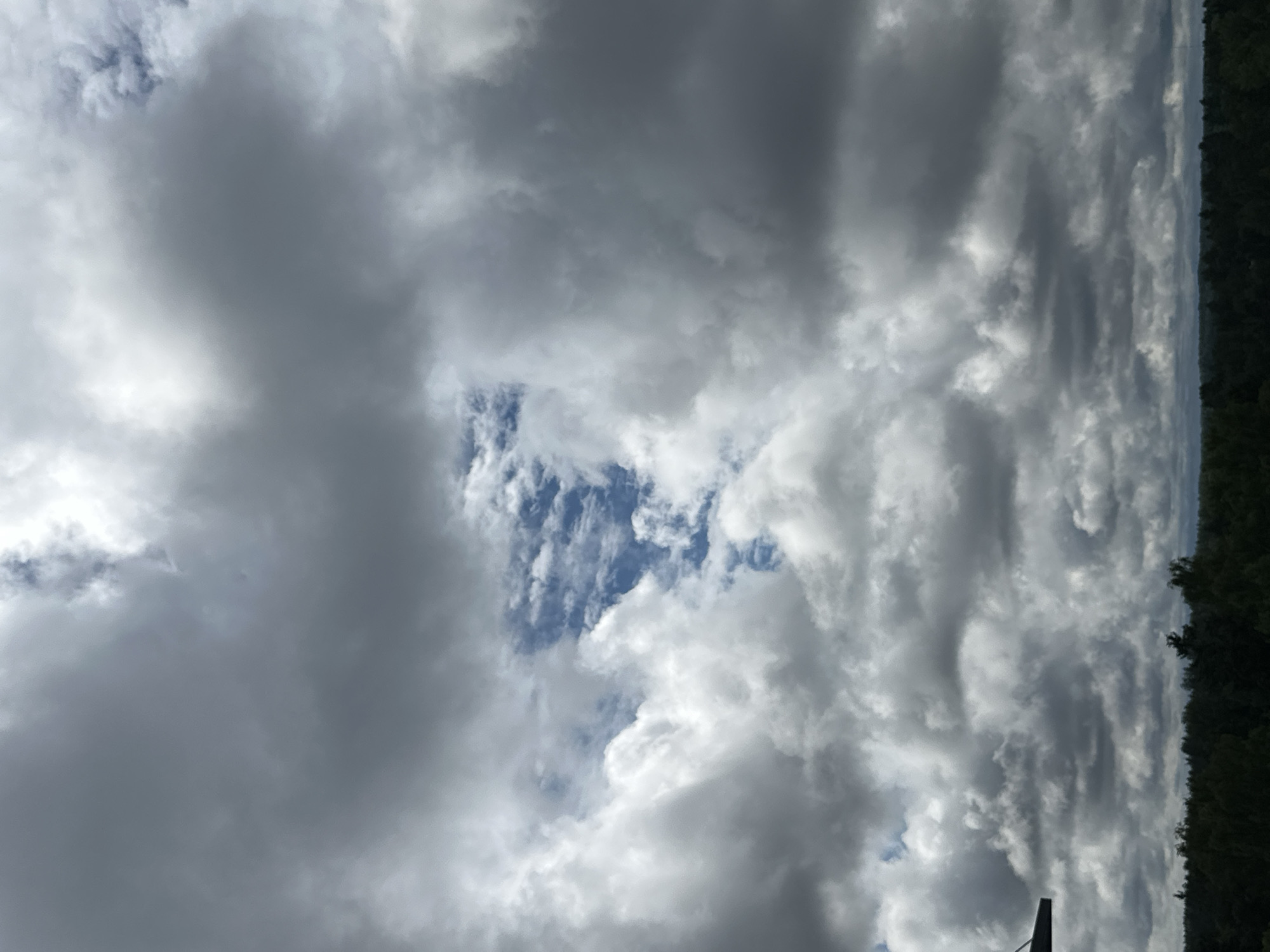 A view from a high vantage point looks out over a dense forest canopy under a dramatic sky filled with dark, heavy clouds. A small patch of blue sky is visible through a break in the clouds, revealing hints of sunlight. In the lower left corner of the frame, a dark, angular object juts into the scene, its purpose and origin unclear. The overall impression is one of an impending storm or the aftermath of one, with the natural landscape beneath a powerful and changing sky.