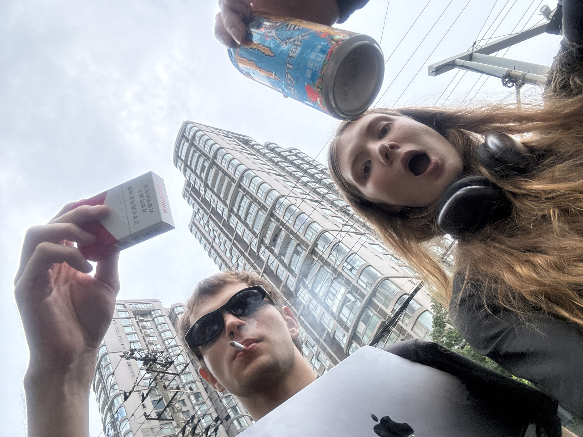 A low-angle, upward-facing urban snapshot captures two individuals in what appears to be the Jing'an District, China, under an overcast sky. In the foreground, a male subject with black sunglasses is smoking a cigarette, holding a silver laptop (likely a MacBook, indicated by the Apple logo) with his right hand. Above his left hand, he holds a red and white packet of cigarettes, on which Chinese health warnings are clearly visible: "尽早戒烟有益健康" (Quit smoking early for health) and "戒烟可减少 对健康的危害" (Quitting smoking can reduce harm to health).
To the top-right, a female subject with long, light brown hair wears black over-ear headphones around her neck and displays a surprised or expressive open-mouthed facial expression. She holds an upside-down blue Monster Energy drink can, with its distinctive logo and text visible, above her head.
The background is dominated by modern, tall high-rise buildings with many windows, characteristic of an urban cityscape. Overhead utility lines and poles are also visible, further establishing the metropolitan setting. The image uses an unconventional perspective, focusing on the subjects and their personal items against the backdrop of the city on a cloudy day.