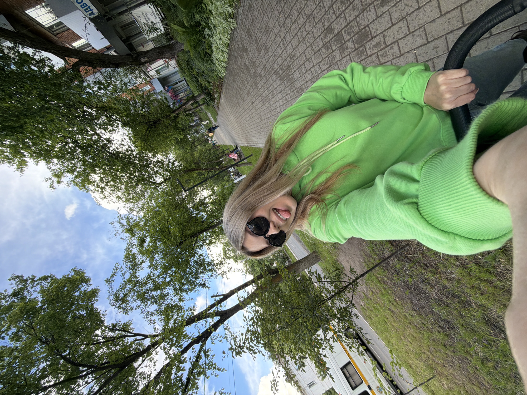 A playful selfie captures a young woman outdoors in Chaikovsky, Russia. The image is taken from a low, tilted angle, showing her looking directly at the camera with her tongue sticking out, suggesting a lighthearted mood. She has long blonde hair and is wearing dark, round sunglasses and a vibrant lime green hooded sweatshirt. Her right hand is visible, resting on a dark, textured handle, possibly from a stroller or similar vehicle. Part of her light blue jeans can also be seen.
The setting appears to be a pedestrian area or park, with a neatly paved path made of grey interlocking bricks extending into the distance. Lush green trees with abundant foliage line the path and frame the upper portion of the scene. Above the trees, a bright blue sky with scattered white clouds indicates a clear, sunny daytime. In the background, behind the trees, several buildings are visible. One prominent building displays a sign with Cyrillic text, including "АВС" and "ЭЛЕКТРИЧЕСКАЯ ЭНЕРГИЯ" (Electric Energy), confirming the Russian context. Further in the background, a white van with yellow stripes and the partial Cyrillic word "ПОЧТ" (likely "ПОЧТА" for Post) is identifiable. The overall impression is one of a cheerful moment captured on a pleasant day.