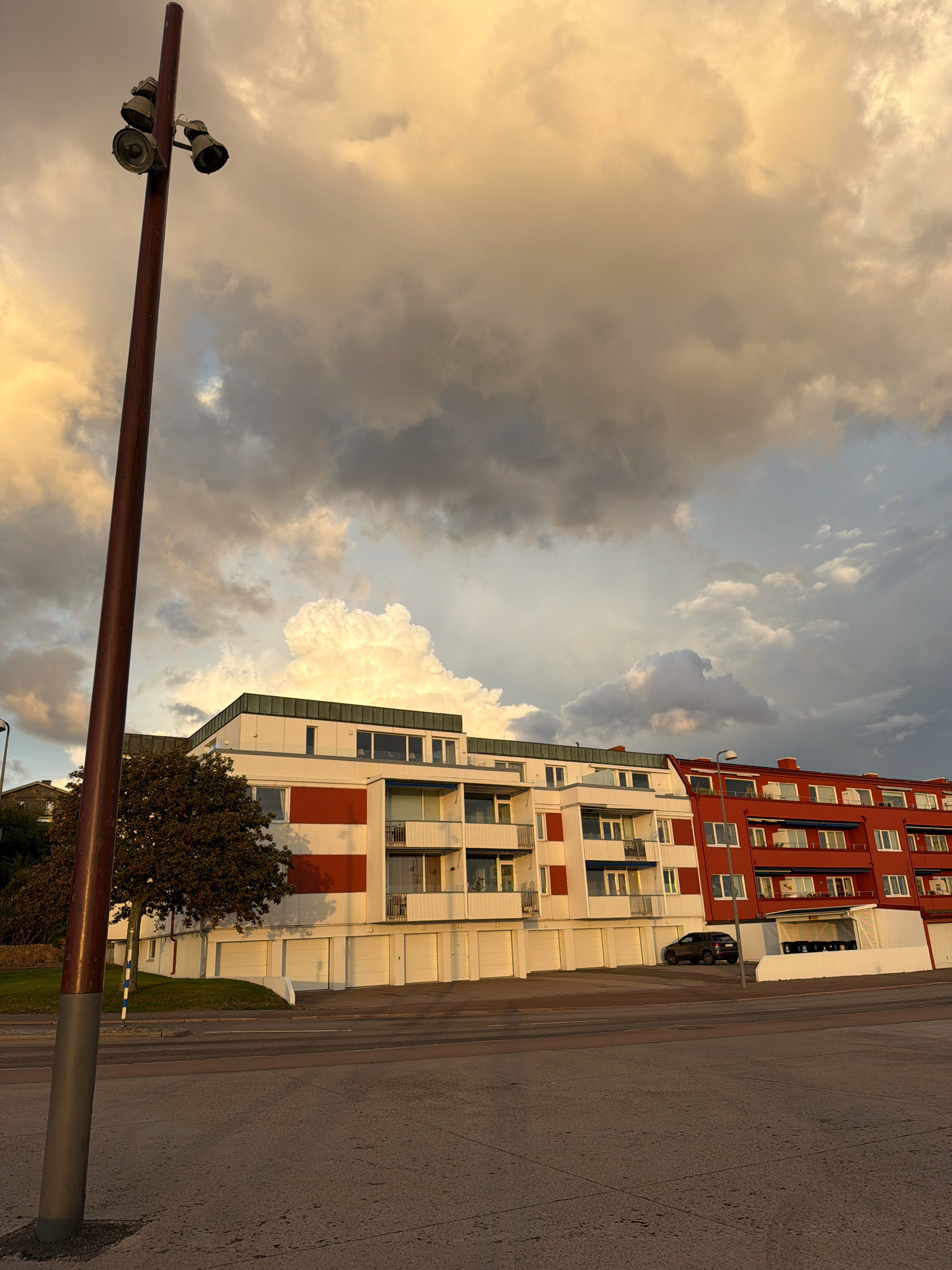 This is a photograph taken in the city of Helsingborg, Sweden, looking towards apartment buildings. The scene is set during what appears to be either sunrise or sunset, indicated by the warm, golden light illuminating the sky and casting long shadows. In the foreground, a tall, dark pole with three lights is positioned to the left. Beyond the pole, two apartment buildings are visible. The building on the left is predominantly white with wide red horizontal bands. It has multiple floors with balconies and windows, and a row of garages at the bottom. A tree with dark foliage is situated in front of this building. The building to the right is painted a bright red and also features several floors with balconies and windows, as well as garages. A dark-colored car is parked in front of the red building. The sky is filled with dramatic clouds, some appearing dark and stormy, while others are brightly lit by the sun. The ground in the foreground is paved, suggesting a street or parking area.