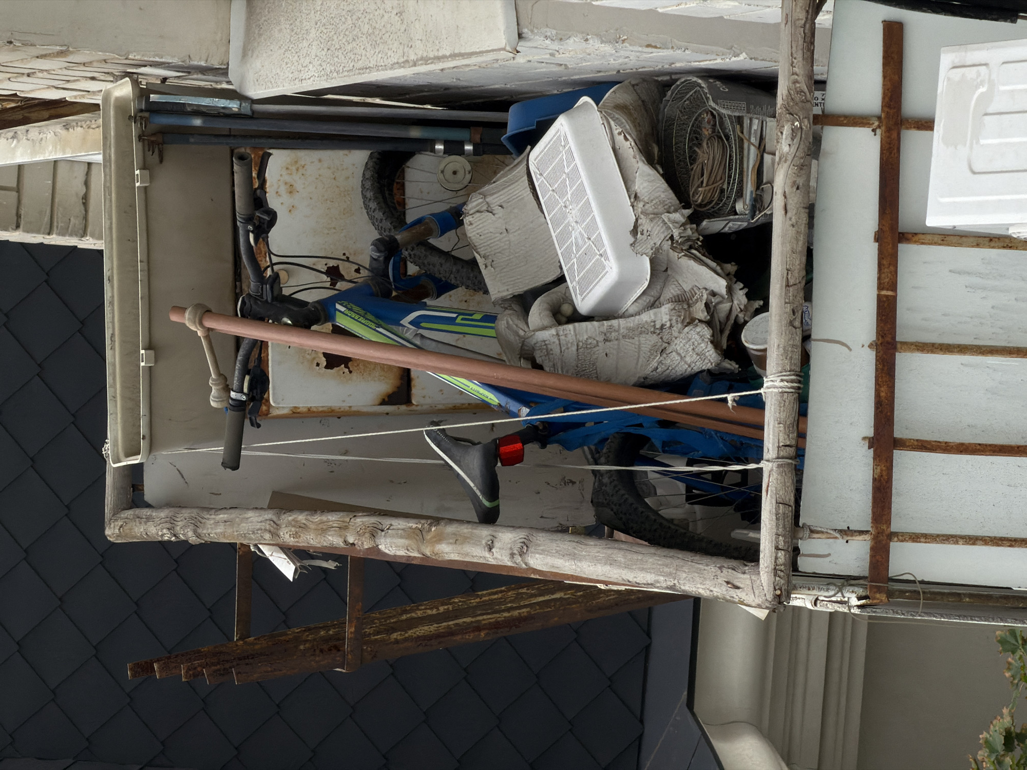 The image depicts a cluttered balcony or outdoor storage area. A blue bicycle with green accents is leaning against a metal structure. Various items are piled around it, including a white plastic tray, cardboard, a metal basket, and what appears to be a rusty metal ladder or frame. The background shows a dark, slate-like roof and a textured brick wall. The scene suggests a disorganized storage space in an urban environment.