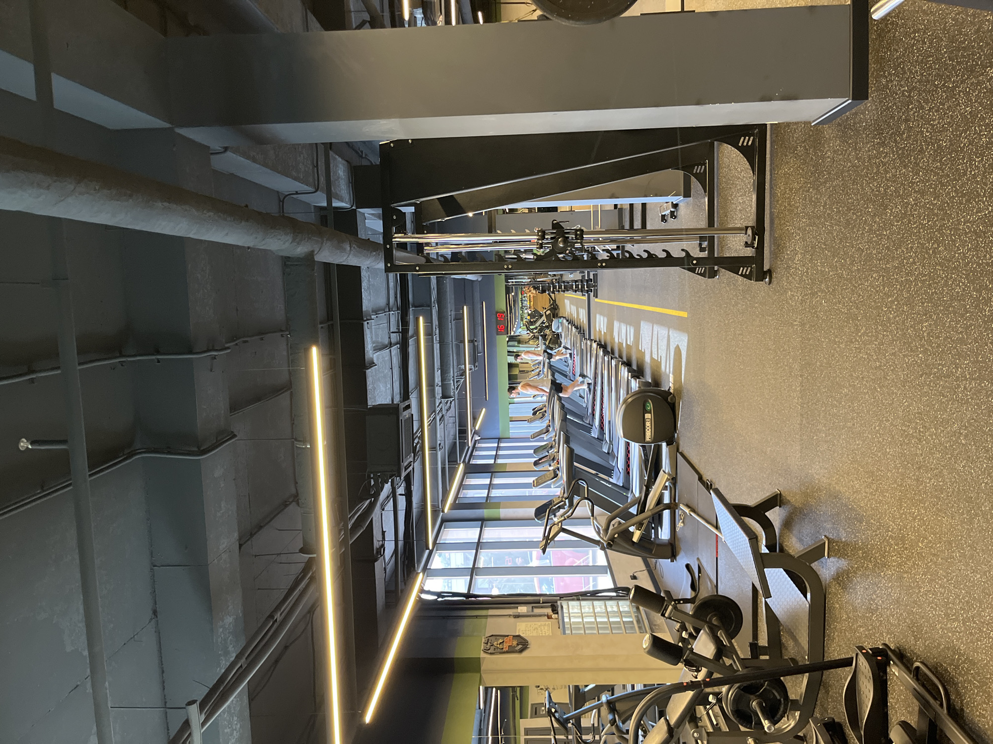 The image shows the interior of a modern gym. In the foreground, there is a black weight bench and exercise equipment. A row of treadmills extends into the background, with one person visible walking on one of them. The gym has a high ceiling with exposed pipes and bright linear lights. Large windows line one wall, allowing natural light to enter and showing a blurred view of the outside. The floor is made of black rubberized material. The gym is located in Sochi, Russia.