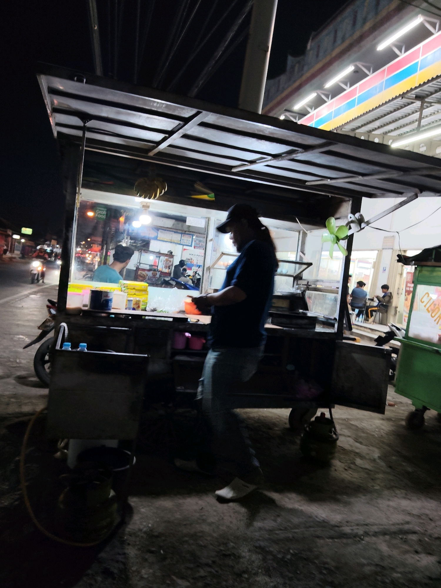 This is a medium-shot, blurry, low-light photograph taken at night on a street in Purwakarta, Indonesia, likely a food stall area. A food cart is prominently featured, with a person in the foreground wearing a dark t-shirt, a baseball cap, and jeans, facing away from the camera and appearing to be working at the stall. The cart itself has a metal frame and is filled with various items, including food containers, bottles, and what appears to be a fan. Behind the cart, through glass, several customers are visible seated at tables, illuminated by overhead lights. Another food stall, painted green, is visible to the right of the frame, and in the background, streetlights and building lights indicate an urban setting. A motorcycle is parked to the left of the food cart. The ground appears to be asphalt with some debris.